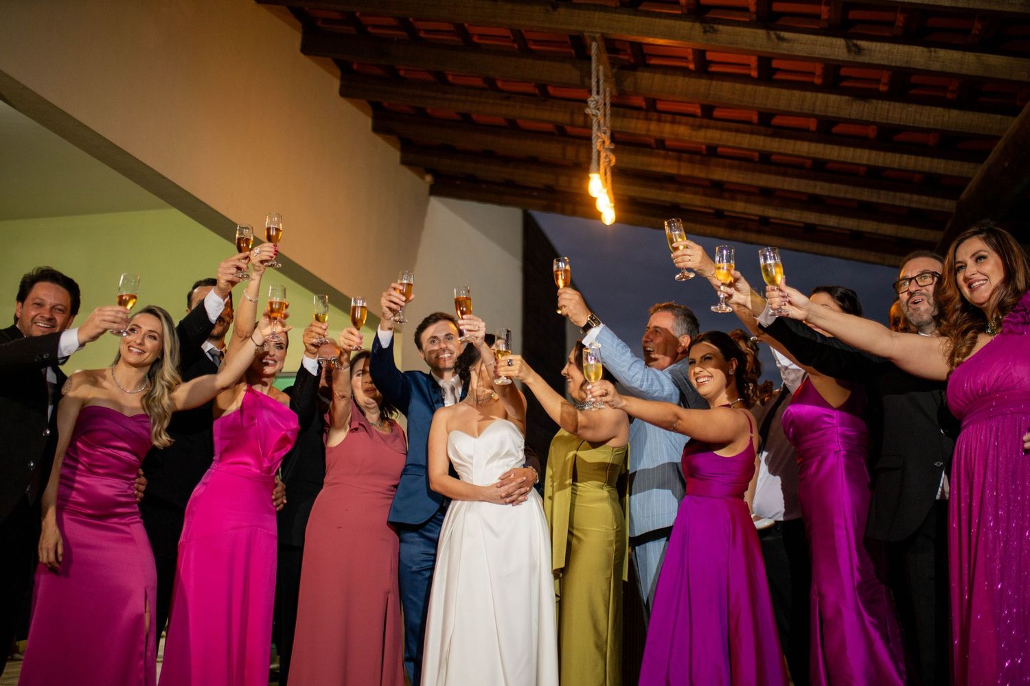 Wedding guests in colorful gowns raise champagne glasses in a joyful toast with bride and groom at elegant reception.