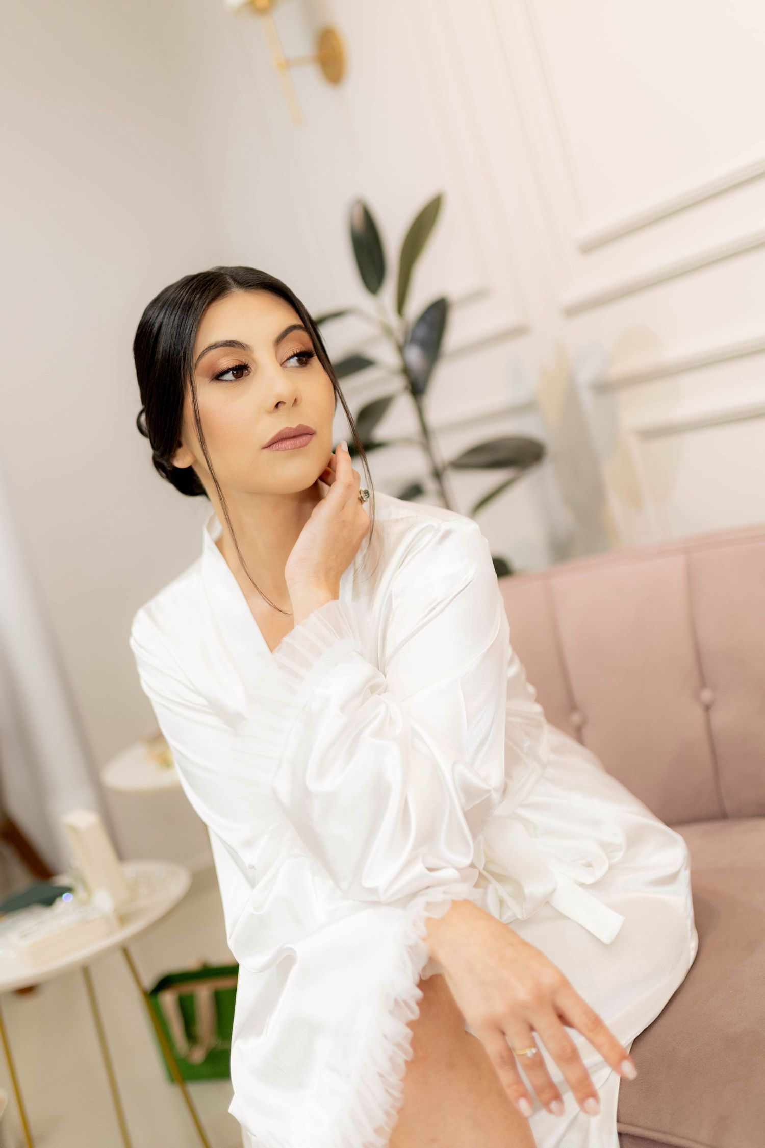 A bride in a white satin robe sits elegantly on a pink chair, touching her chin, with soft natural light filling the room.