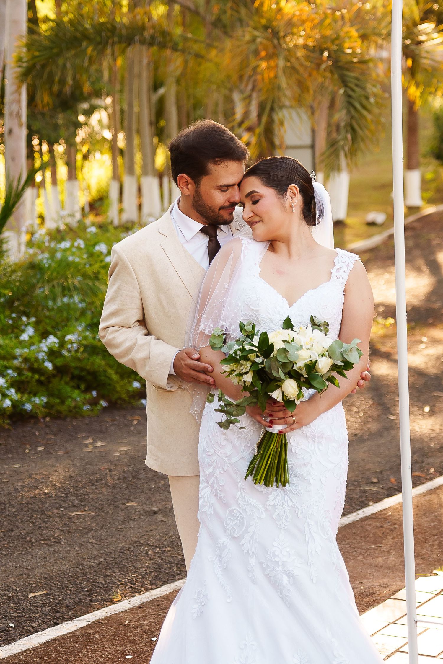Wedding couple touching foreheads tenderly outdoors, bride in lace gown holding white floral bouquet.