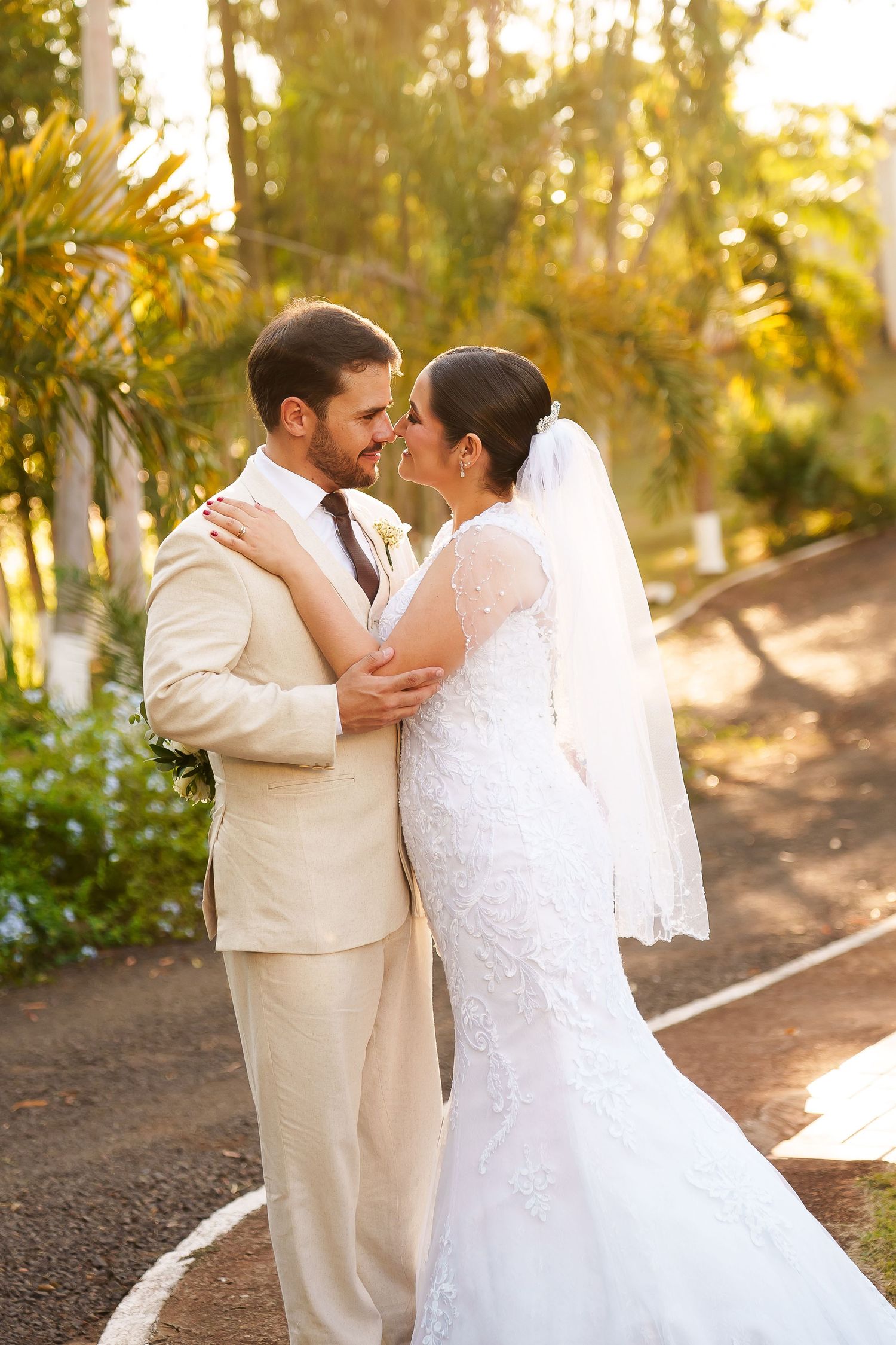 Bride in lace gown and veil embraces groom in beige suit, sharing a romantic moment outdoors with tropical greenery.