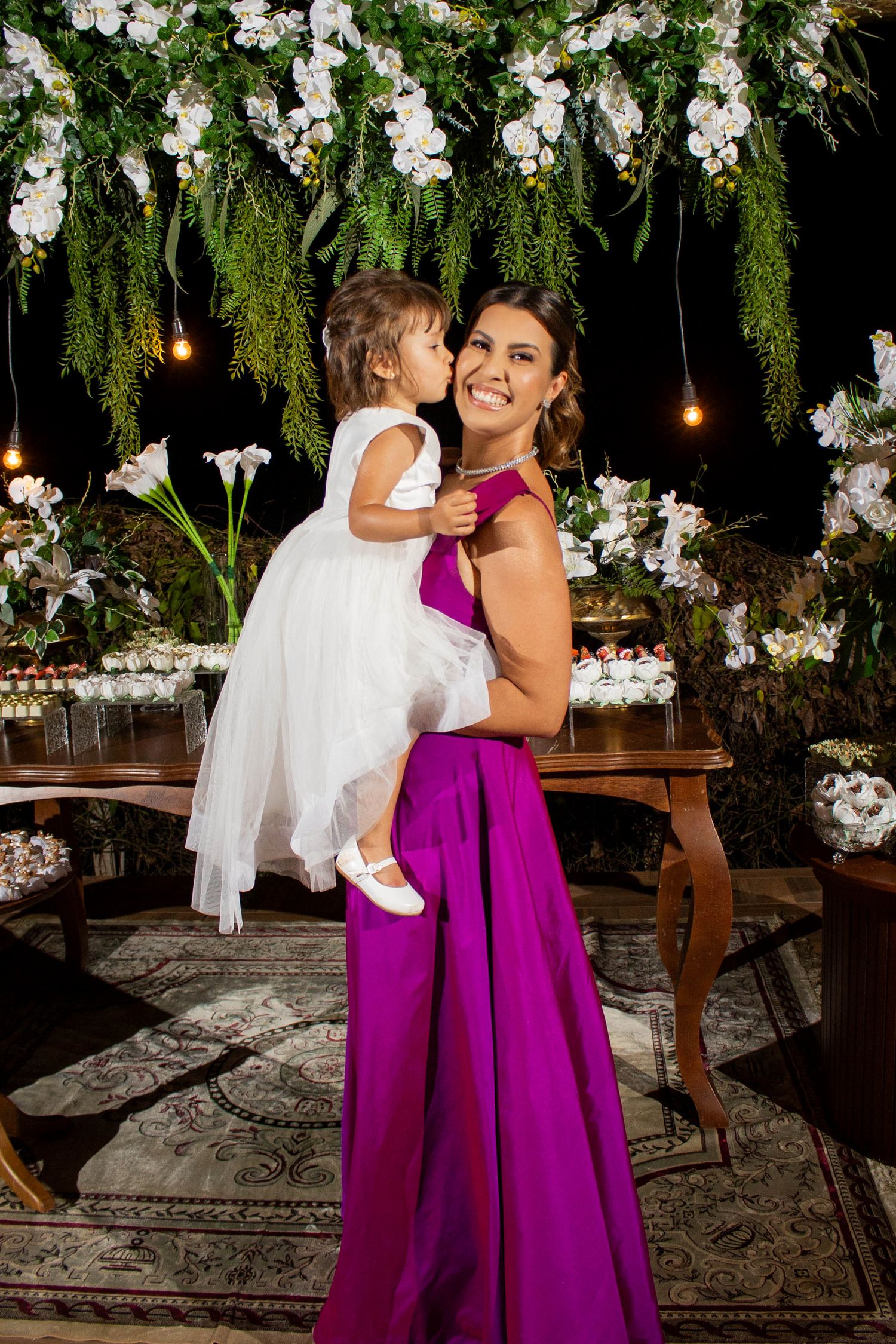 Woman in purple gown holds young girl in white dress at floral decorated event.