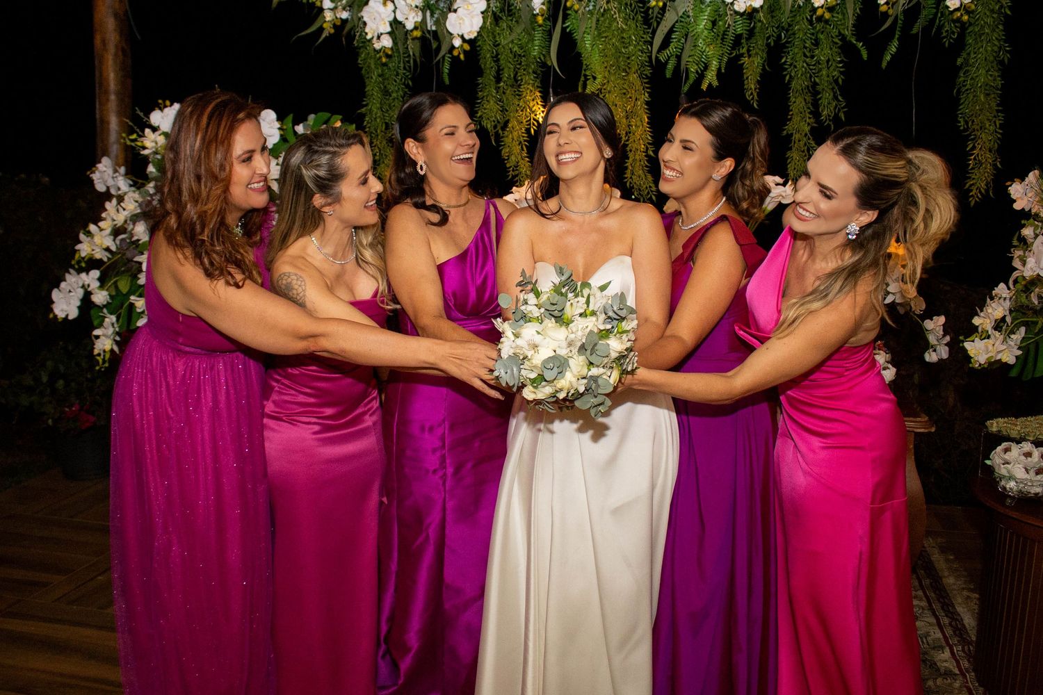 Bride in white gown holds bouquet surrounded by five bridesmaids in pink and purple dresses at outdoor wedding.