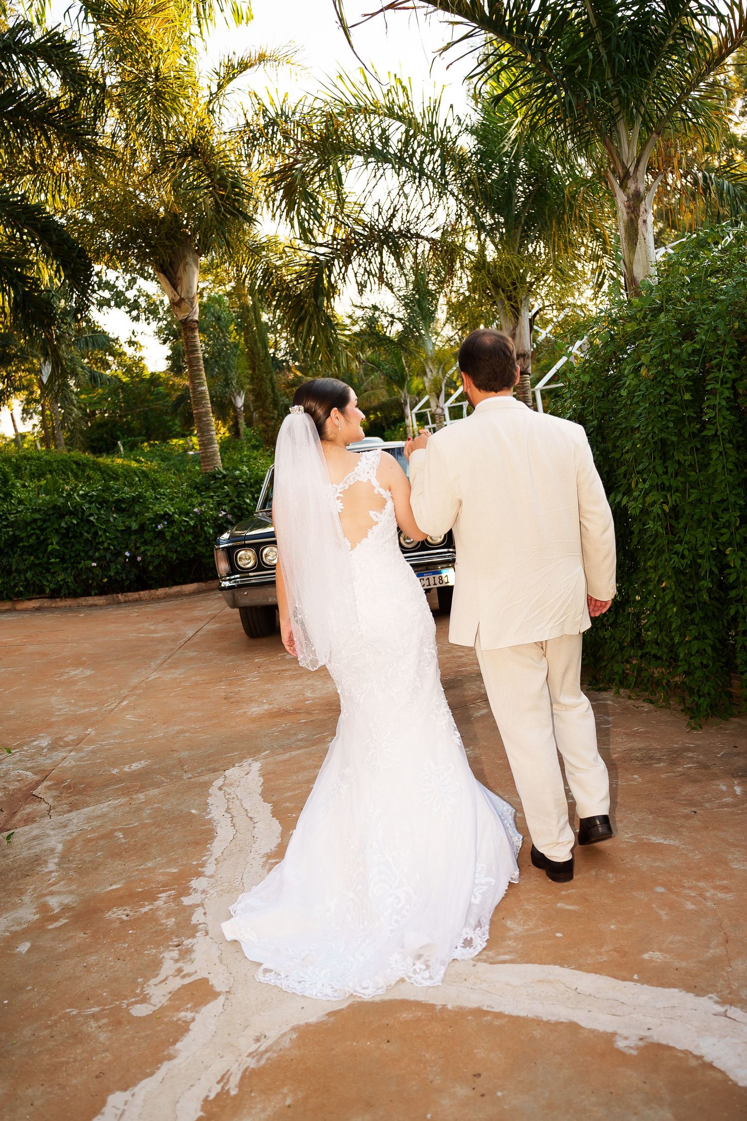 Bride in white lace gown and groom in beige suit walk toward vintage car surrounded by tropical palms.