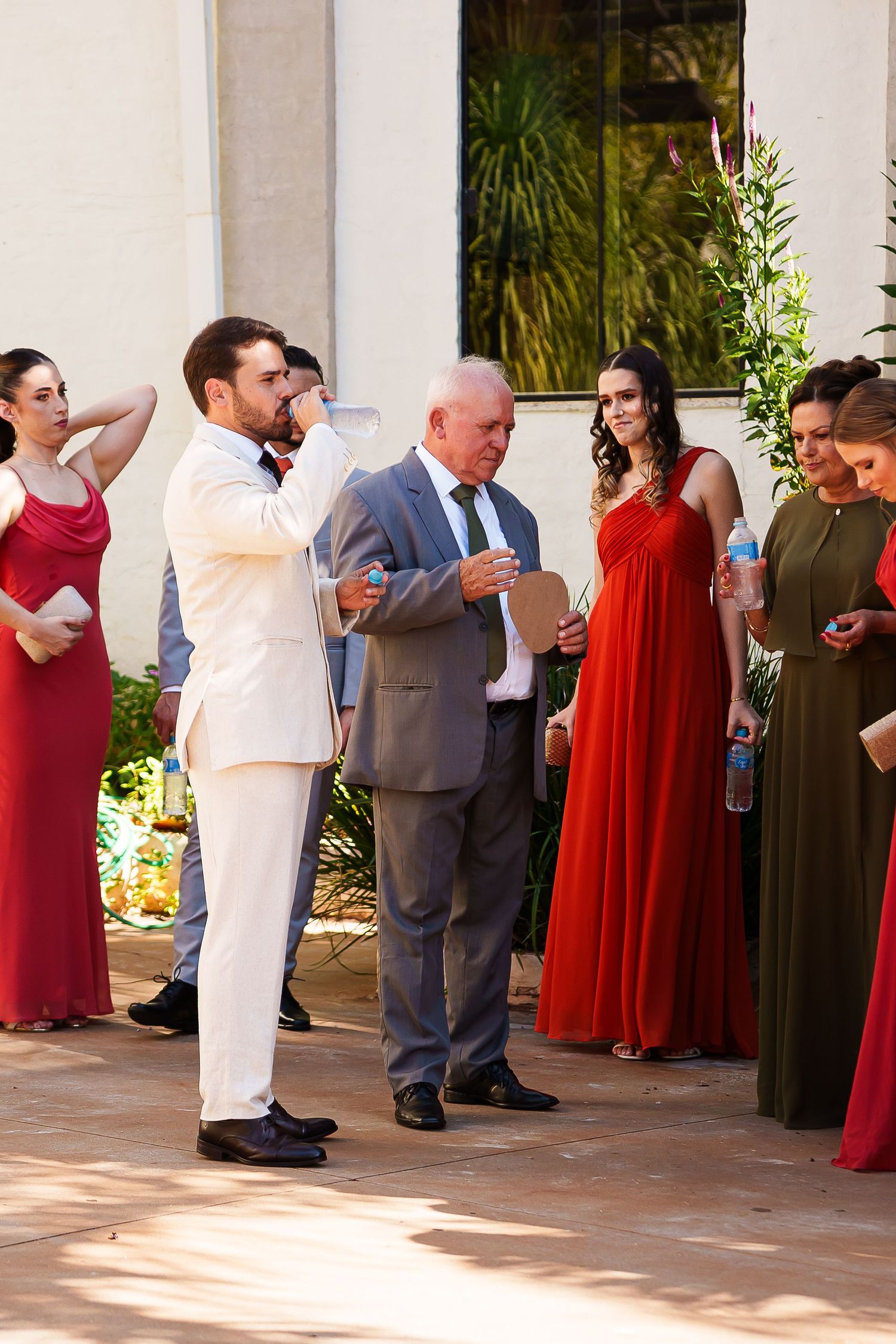 Groom in white suit toasting at outdoor wedding reception with guests in red and green formal attire.