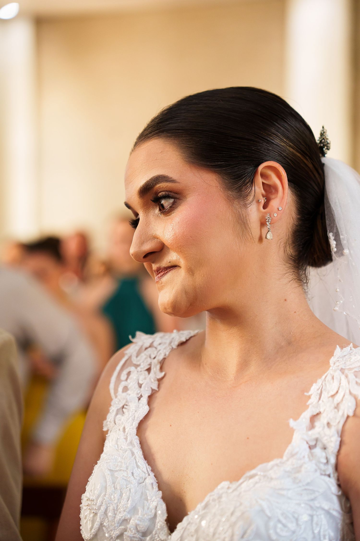 A bride in a white lace gown and veil smiles softly, wearing elegant drop earrings at her wedding reception.
