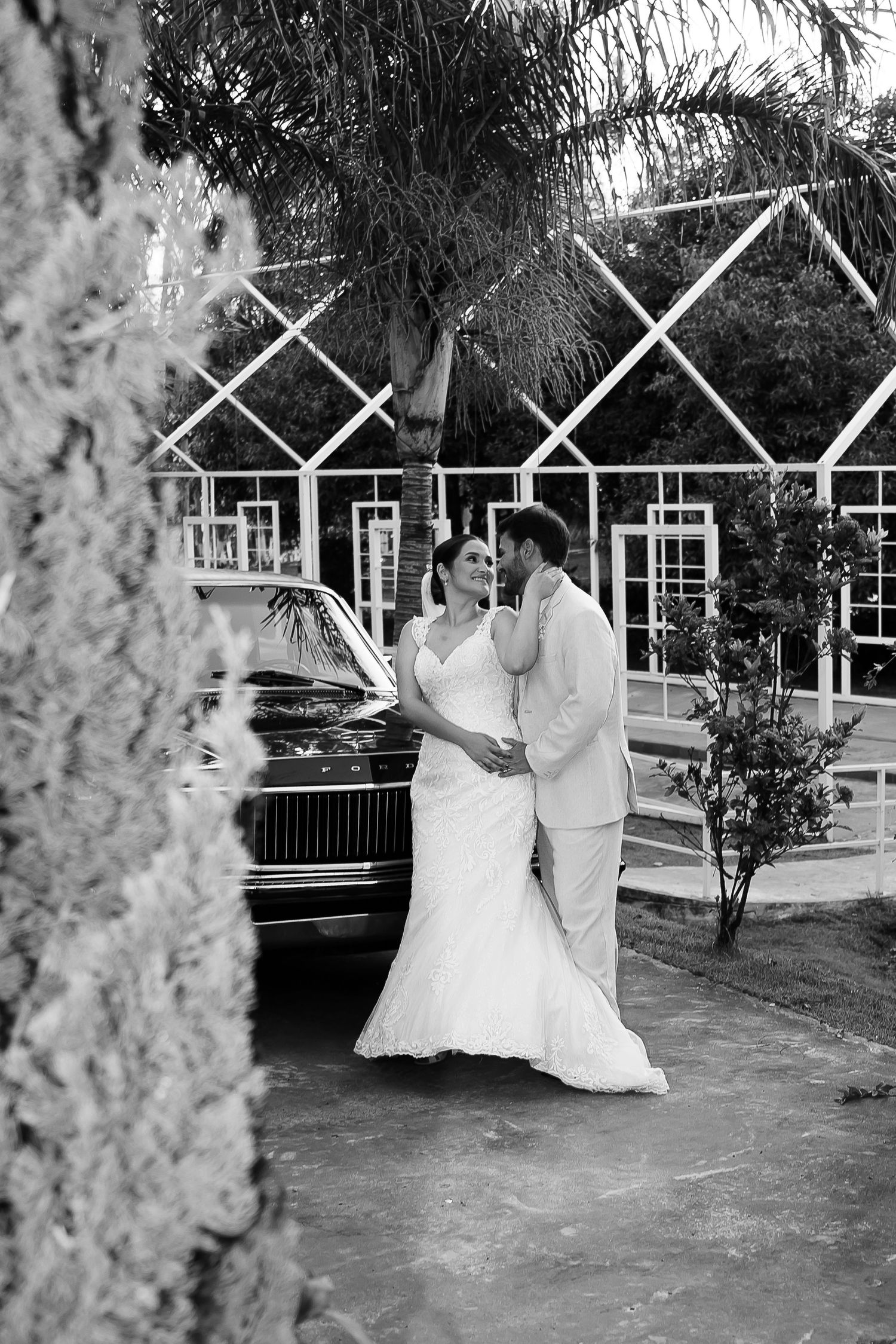 Black and white wedding photo of bride and groom embracing near a classic car with palm trees and a white building behind them.