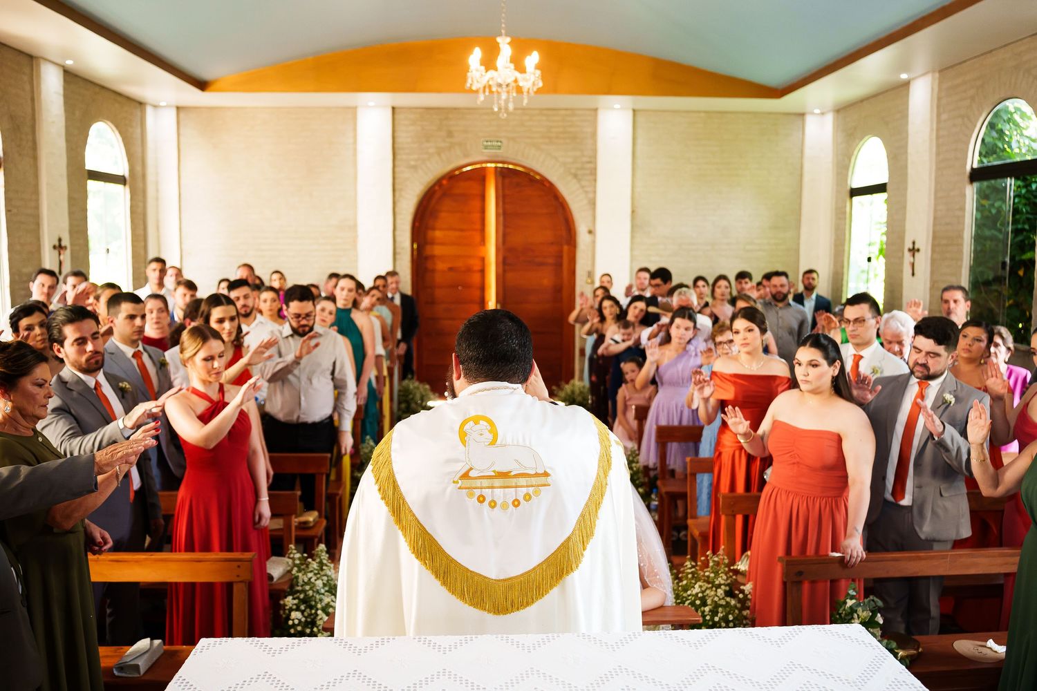 Priest in white vestments leads a wedding ceremony at the altar with guests standing in a small decorated chapel.