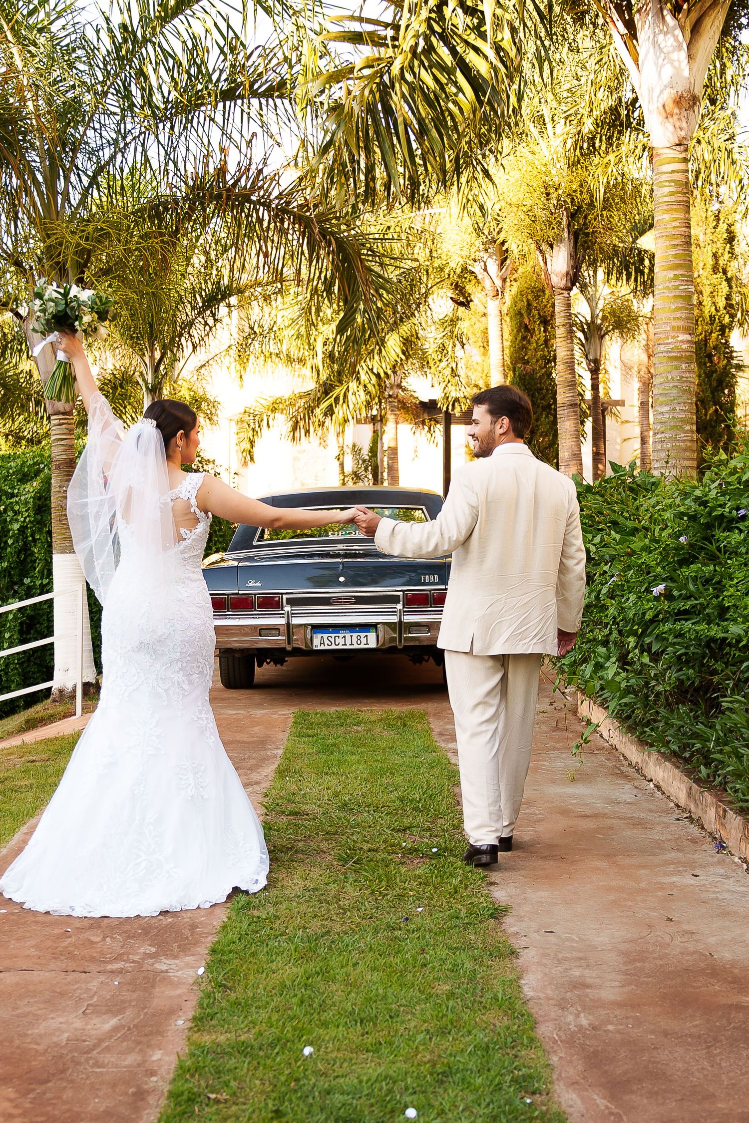 Bride and groom walking hand-in-hand toward a vintage car surrounded by palm trees on their wedding day.