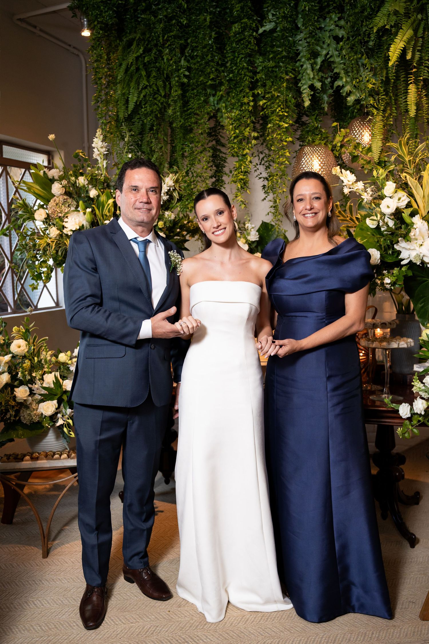 Bride in white strapless gown poses with two guests in navy attire before lush floral greenery backdrop.