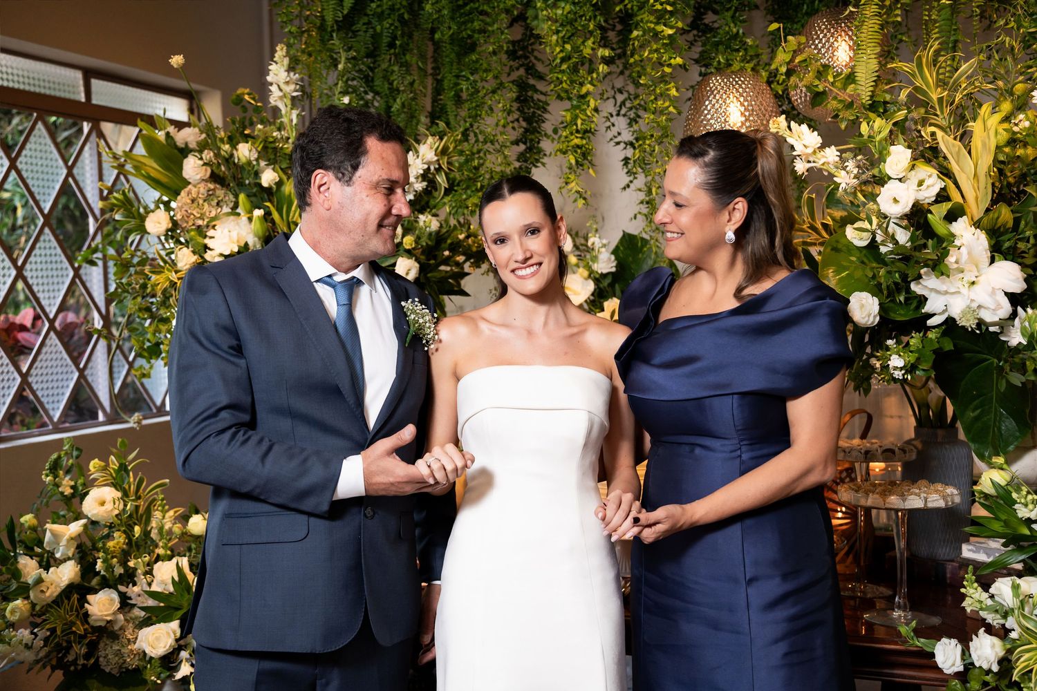 Bride in white strapless gown stands with man in navy suit and woman in blue dress at floral wedding ceremony.