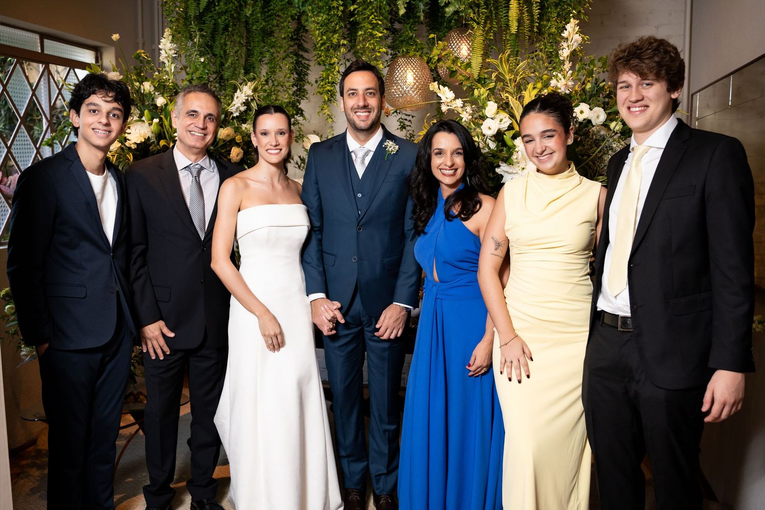 Wedding party of seven posing together, bride in white gown, groom in blue suit, guests in formal attire, floral backdrop.
