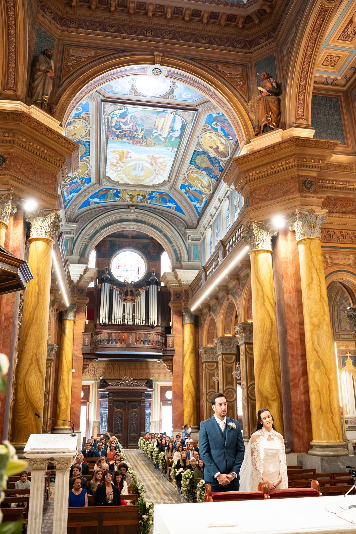 Couple stands at the altar of an ornate church with colorful frescoes, marble columns, and a pipe organ during wedding.