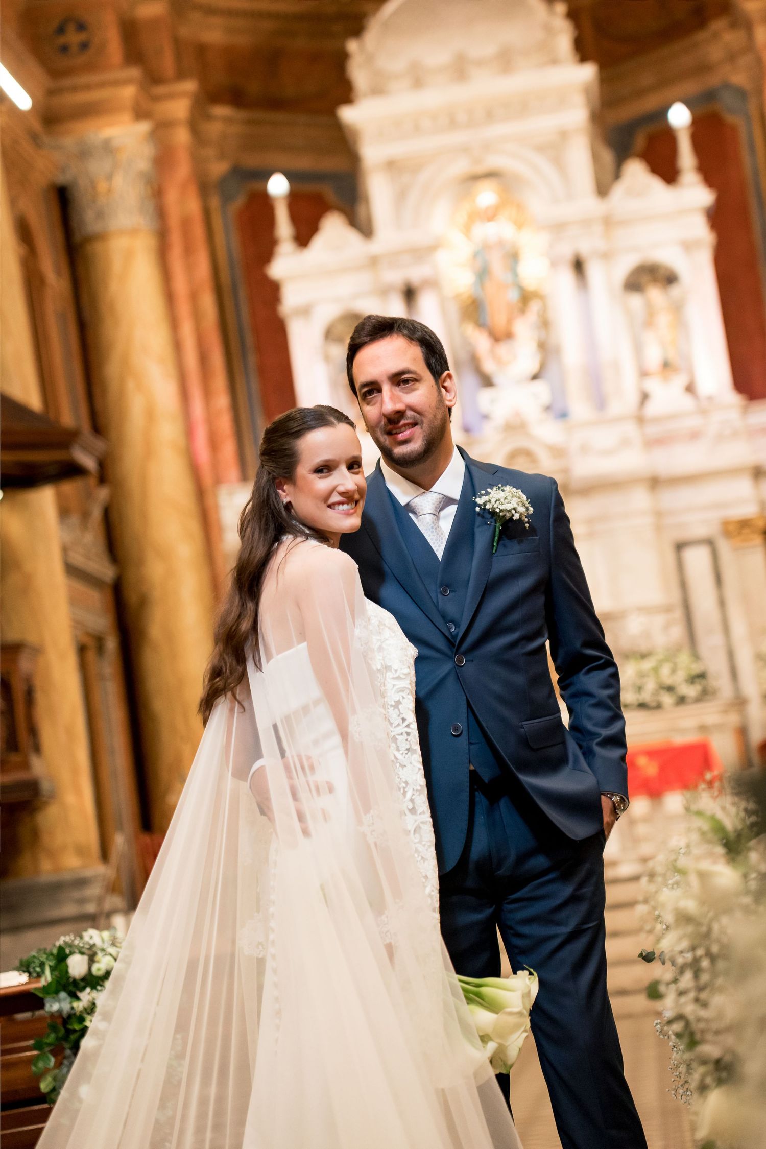 Bride and groom posing together inside an ornate church, smiling, with a grand altar in the background.