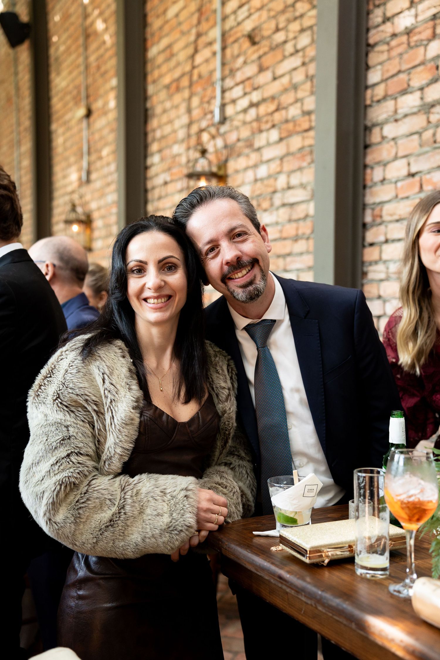 A smiling couple poses together at an elegant event, woman in fur jacket and man in suit with tie, brick wall background.