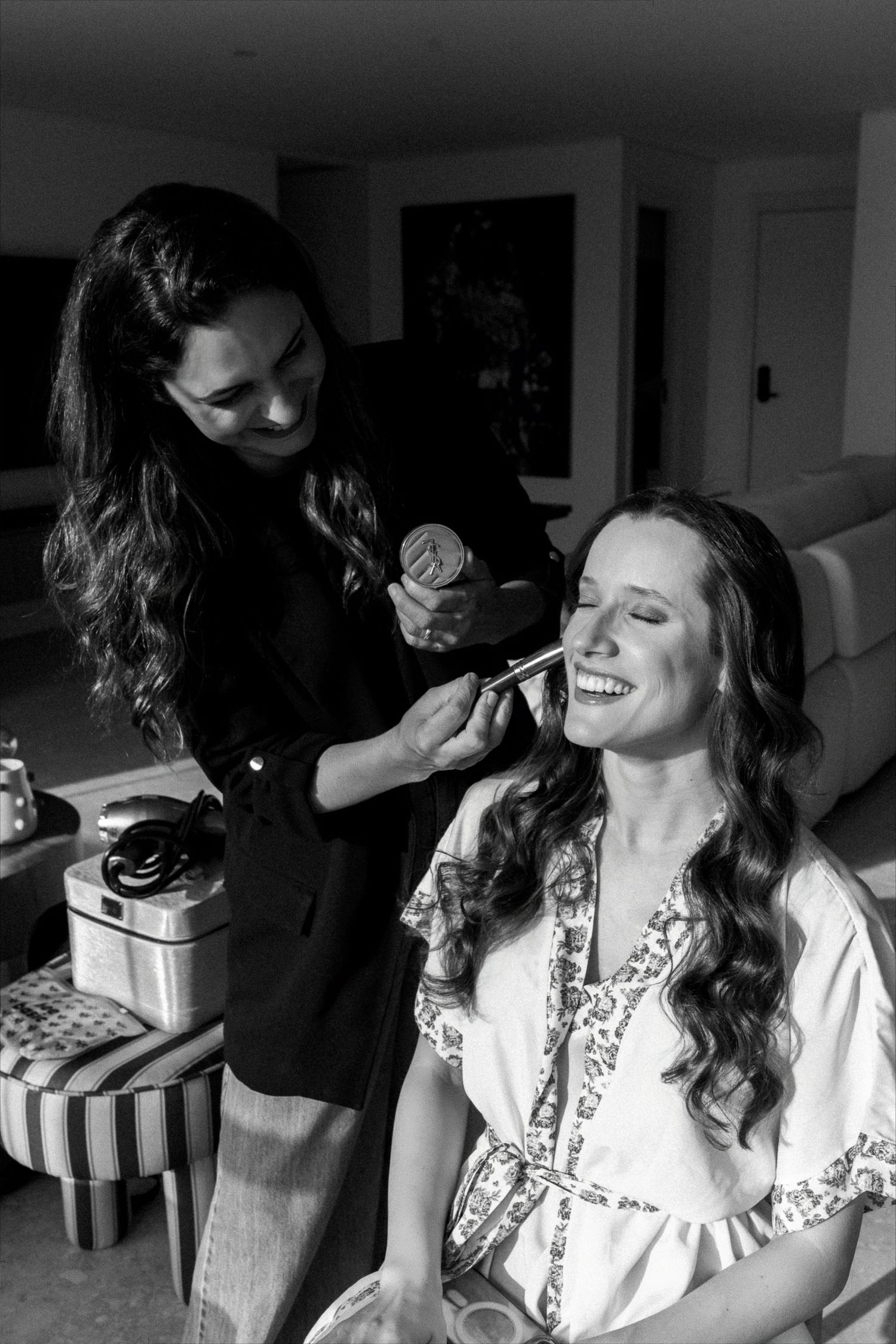 Black and white photo of a makeup artist applying makeup to a smiling bride in a floral robe during wedding preparations.