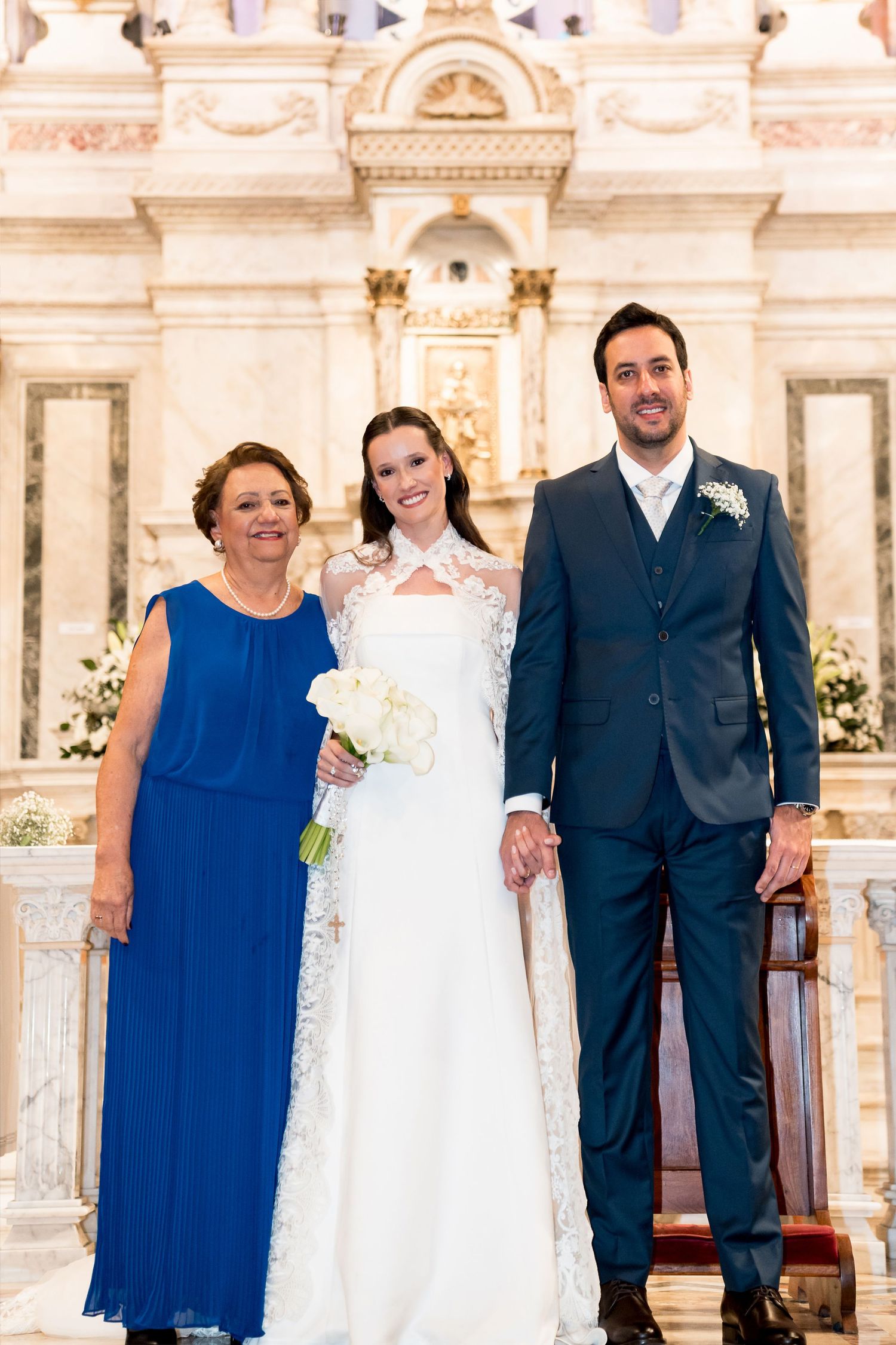 Bride in white lace gown holds bouquet beside groom in navy suit and older woman in blue dress at church altar.