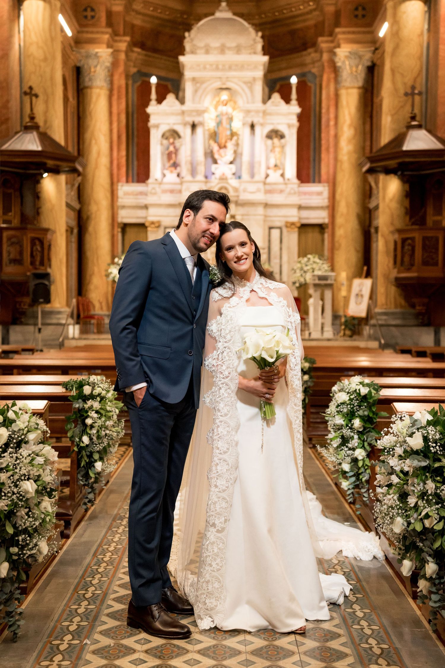 A bride and groom pose together at the altar of an ornate church, she holding a white bouquet.