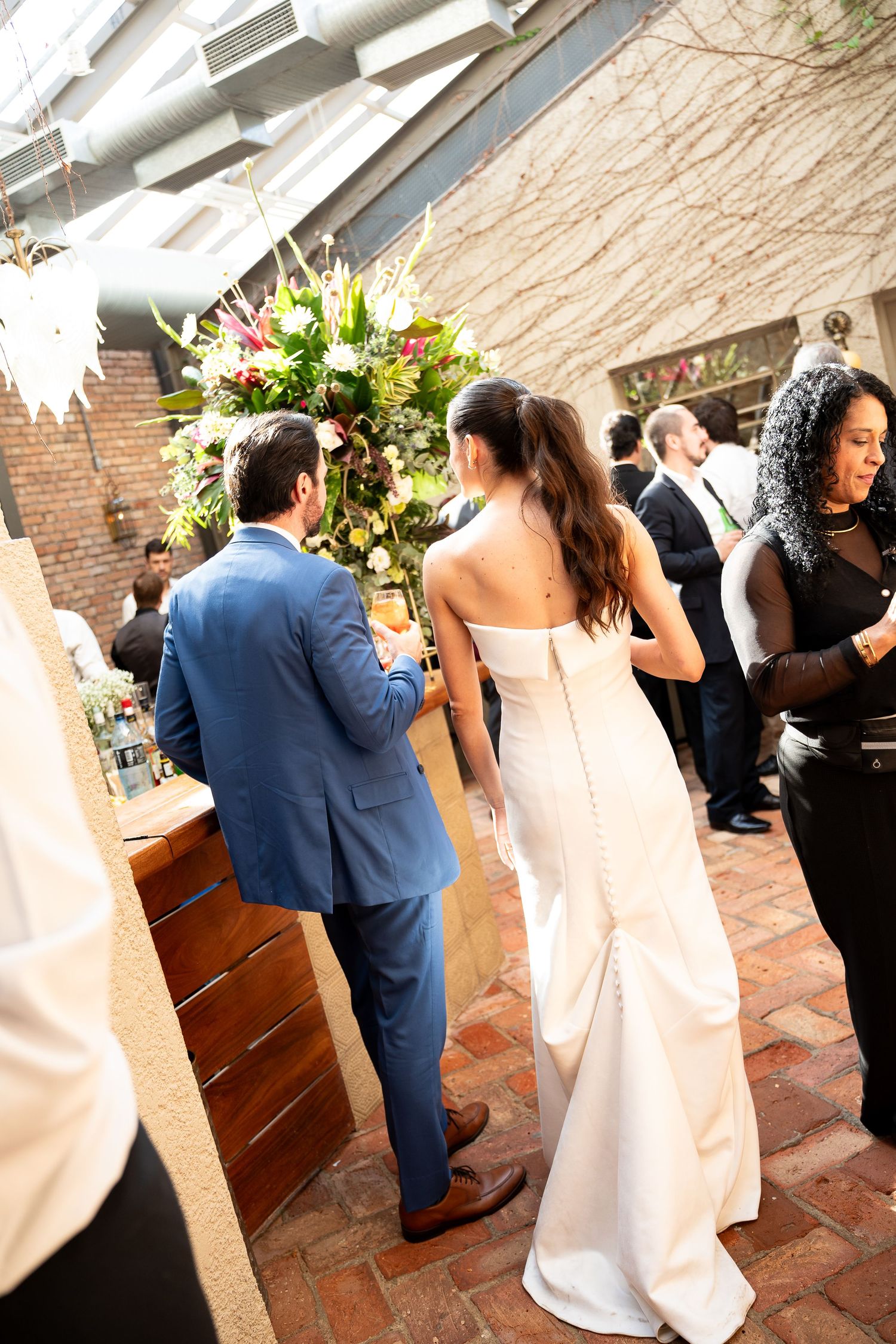 Bride in white strapless gown and groom in blue suit stand at floral altar during indoor wedding ceremony.