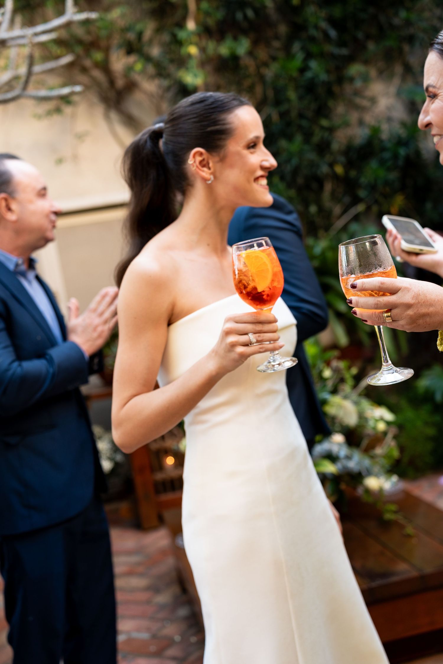 Bride in white strapless gown holding an Aperol spritz at an outdoor wedding reception.