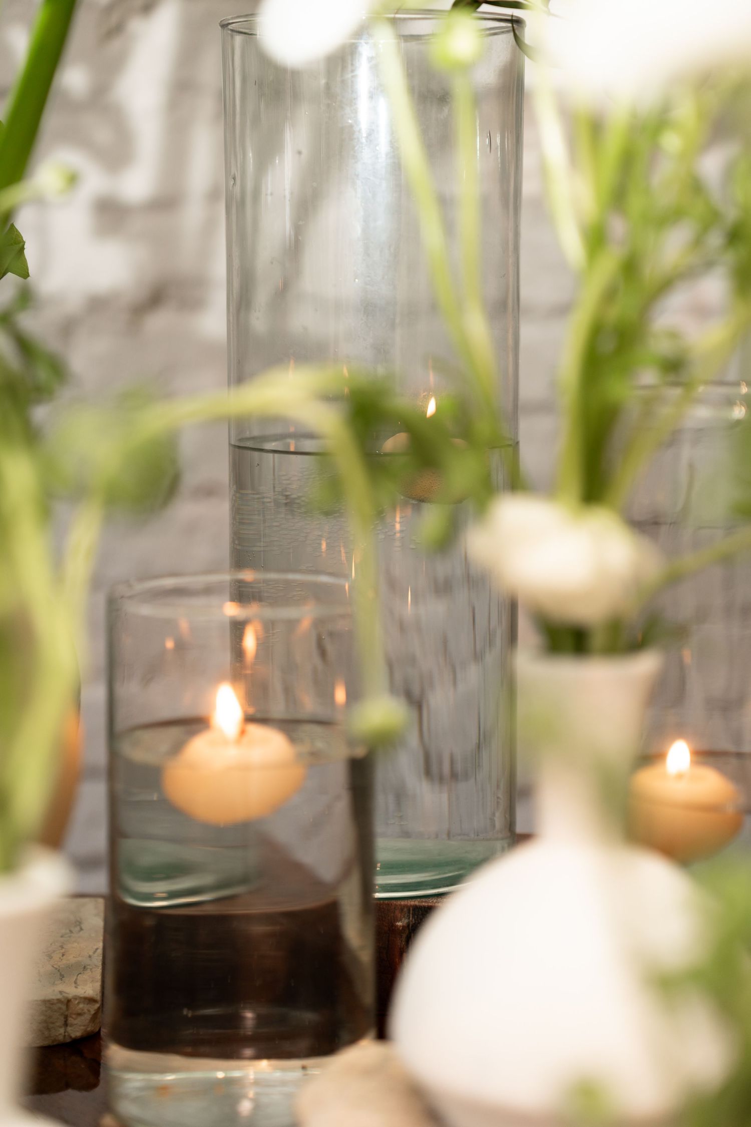Elegant table decor with floating candles in glass cylinders and white flowers in vases against a white brick wall.