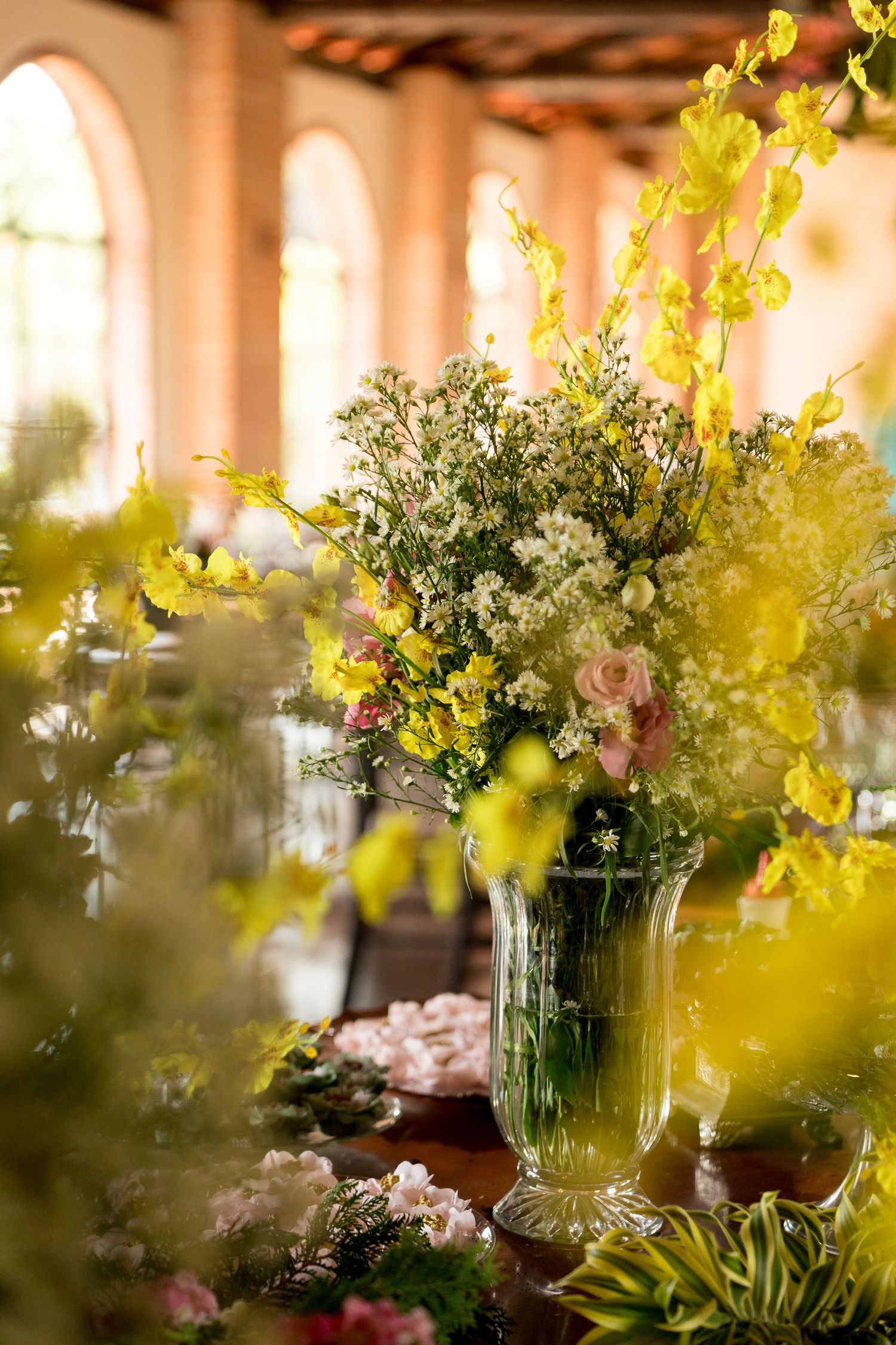 Elegant glass vase with yellow and white floral arrangement on wooden table in a sunlit venue.