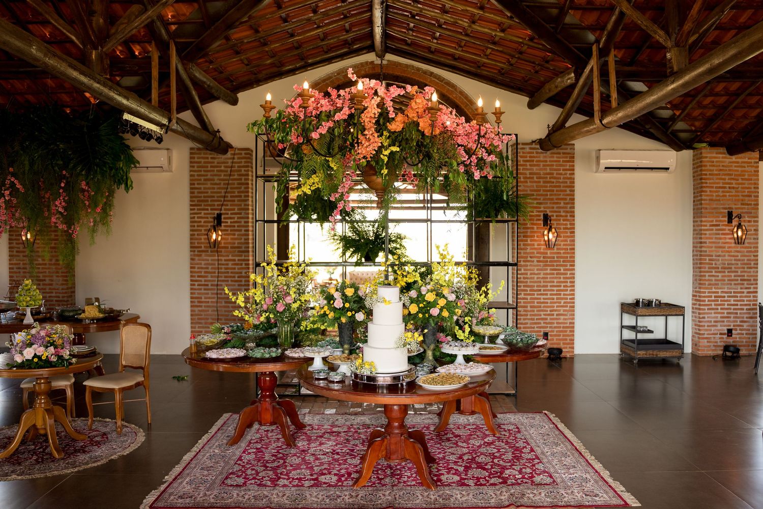Elegant wedding reception hall with floral chandelier, white cake on wooden table, brick walls, and Persian rug decor.