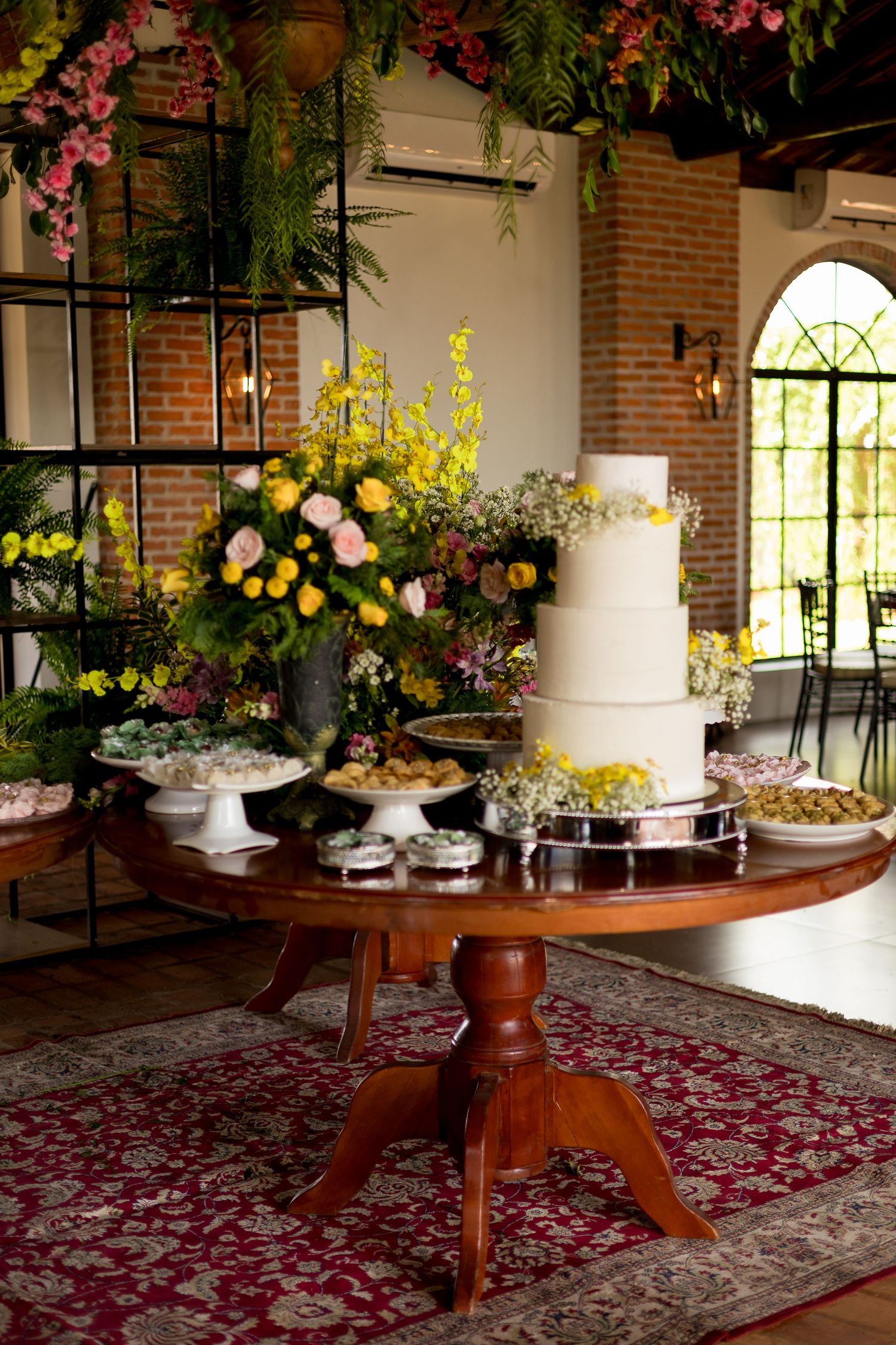Elegant wedding dessert table with white tiered cake, colorful floral arrangements, and assorted sweets on a wooden pedestal table.