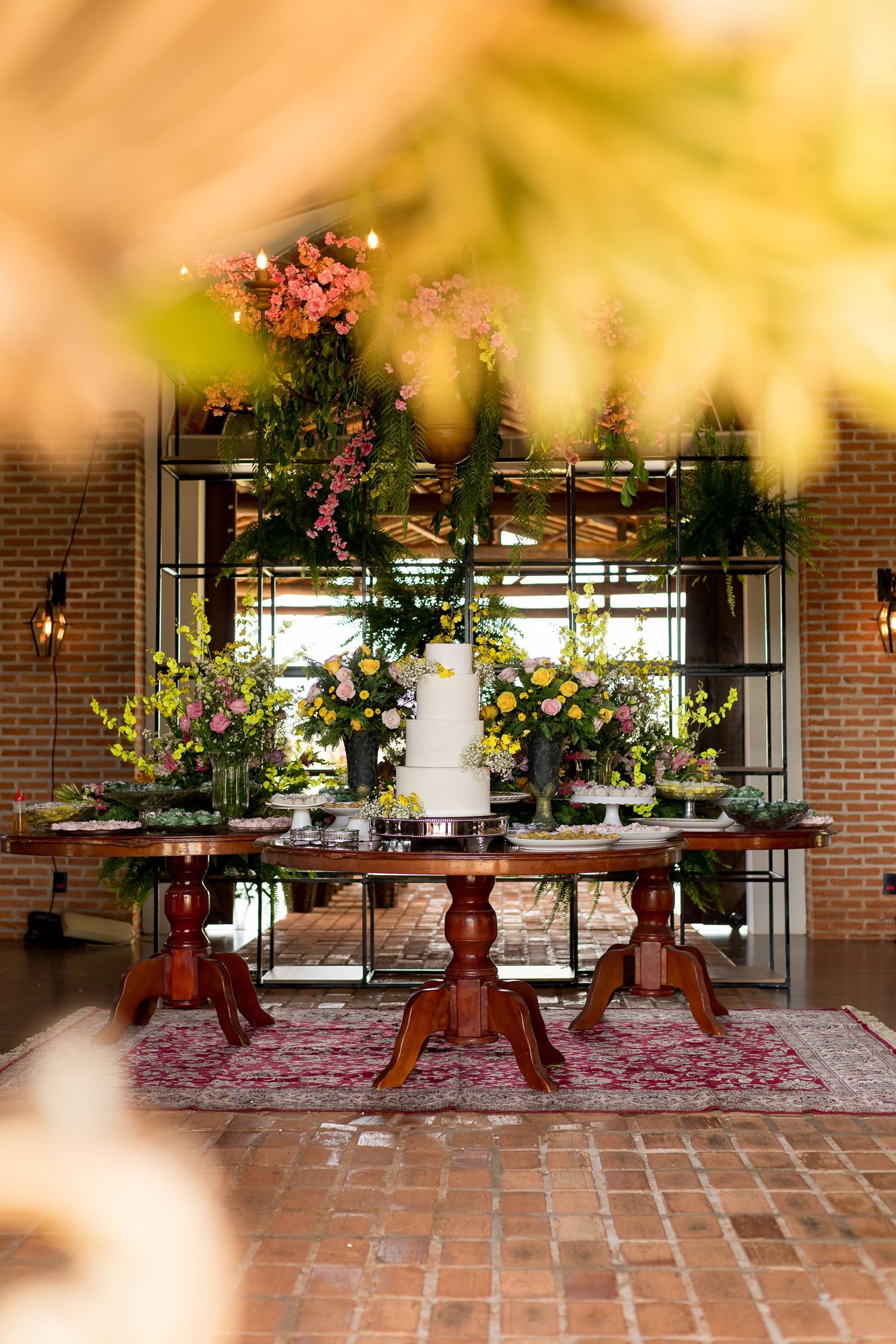 Elegant wedding cake display on rustic wooden tables with lush floral arrangements and brick wall backdrop.