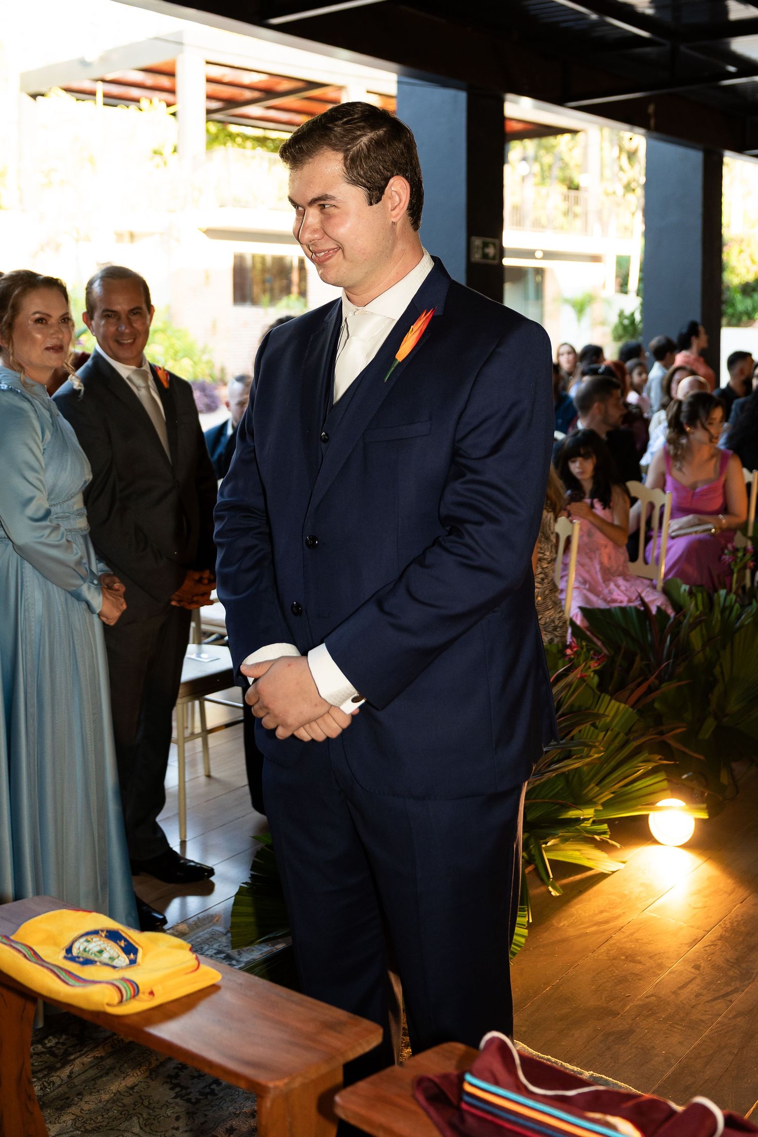 Groom in navy blue suit with orange boutonniere stands at wedding altar, guests seated in background.