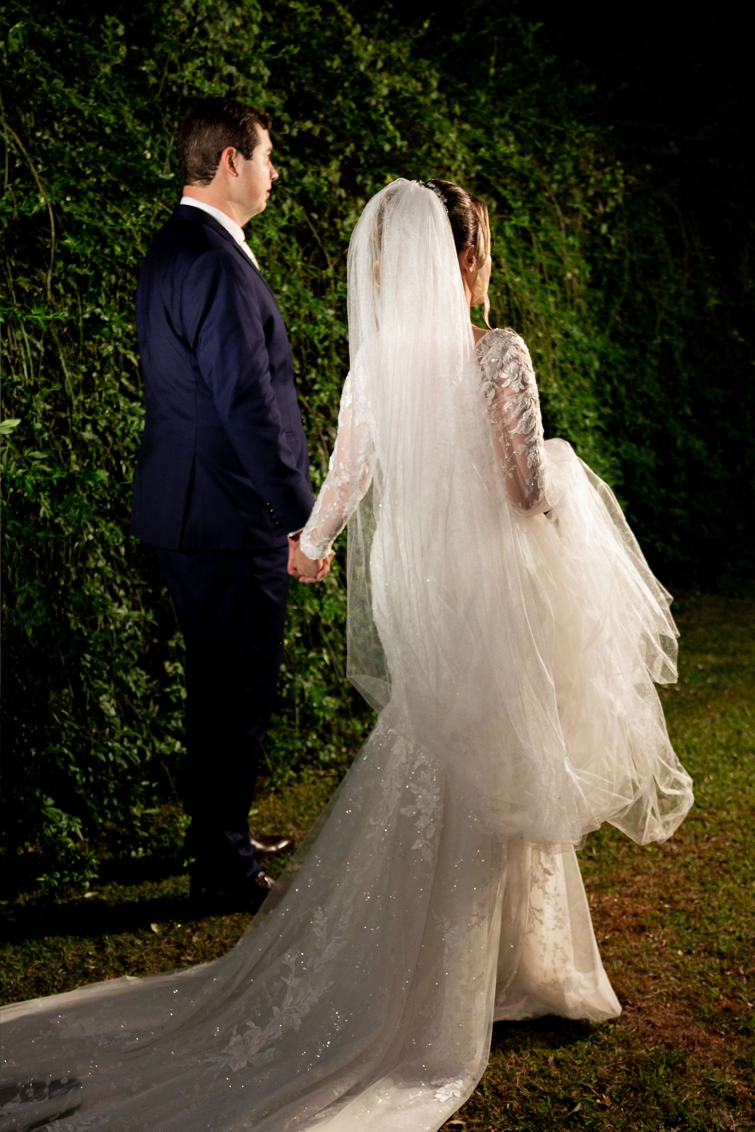 Bride in lace gown with long veil holds hands with groom in navy suit, standing before lush green foliage at night.