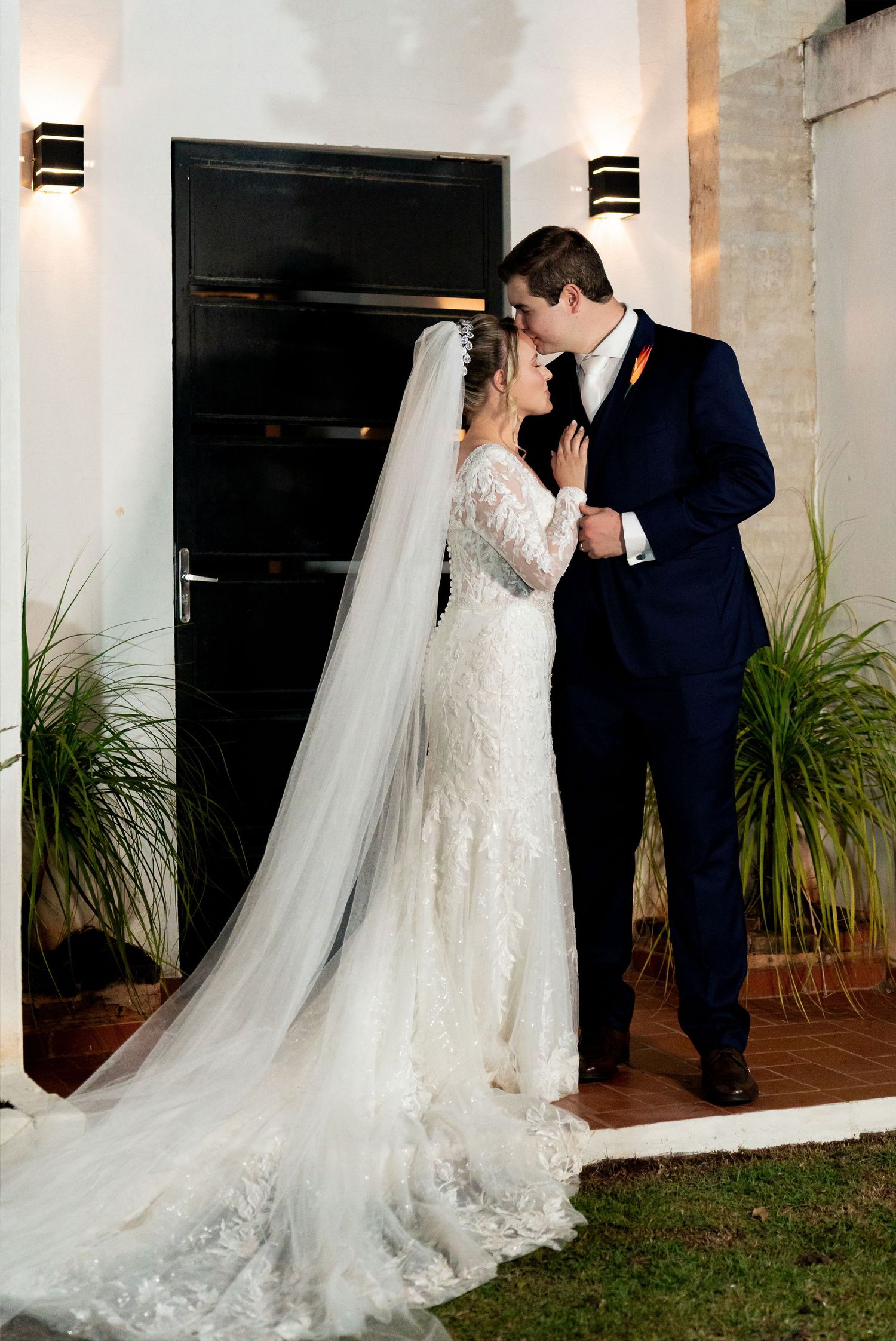 Bride in lace gown with long veil kisses groom in navy suit outside elegant venue doors at night.