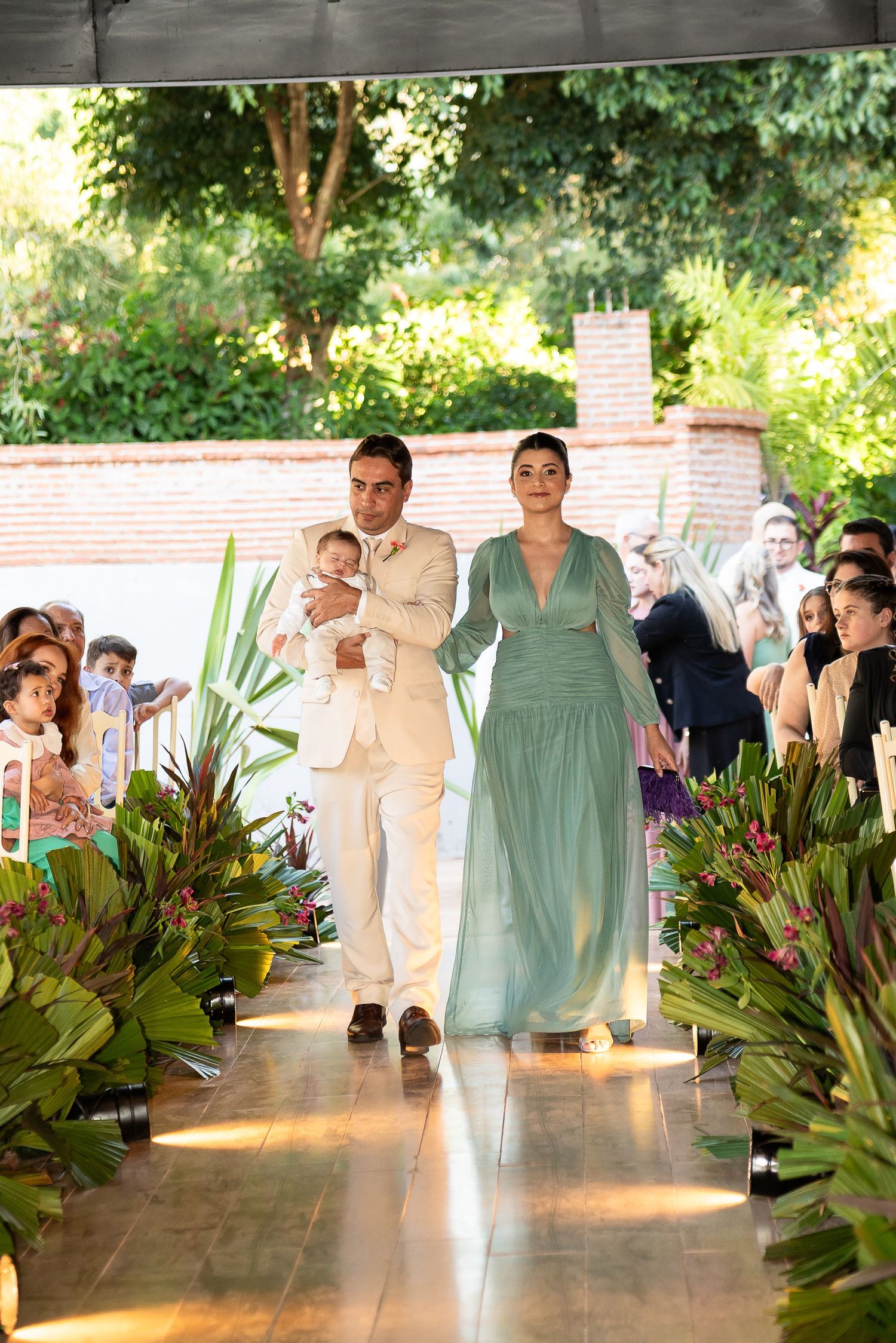 Couple walks down aisle at outdoor wedding, man in cream suit holds baby, woman wears elegant sage green gown.
