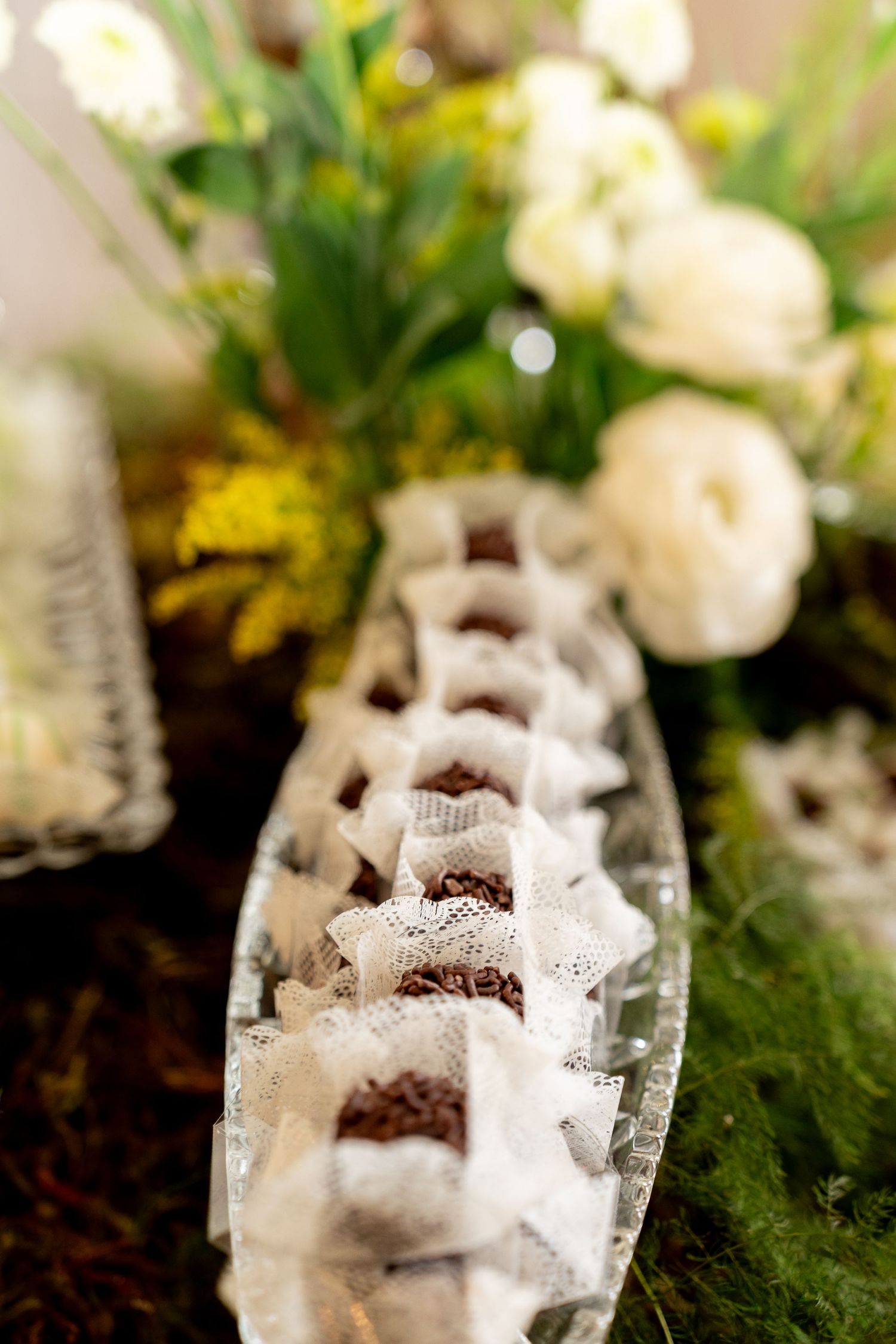 Elegant chocolate truffles in lace wrappers displayed on a glass tray surrounded by white and yellow floral arrangements.