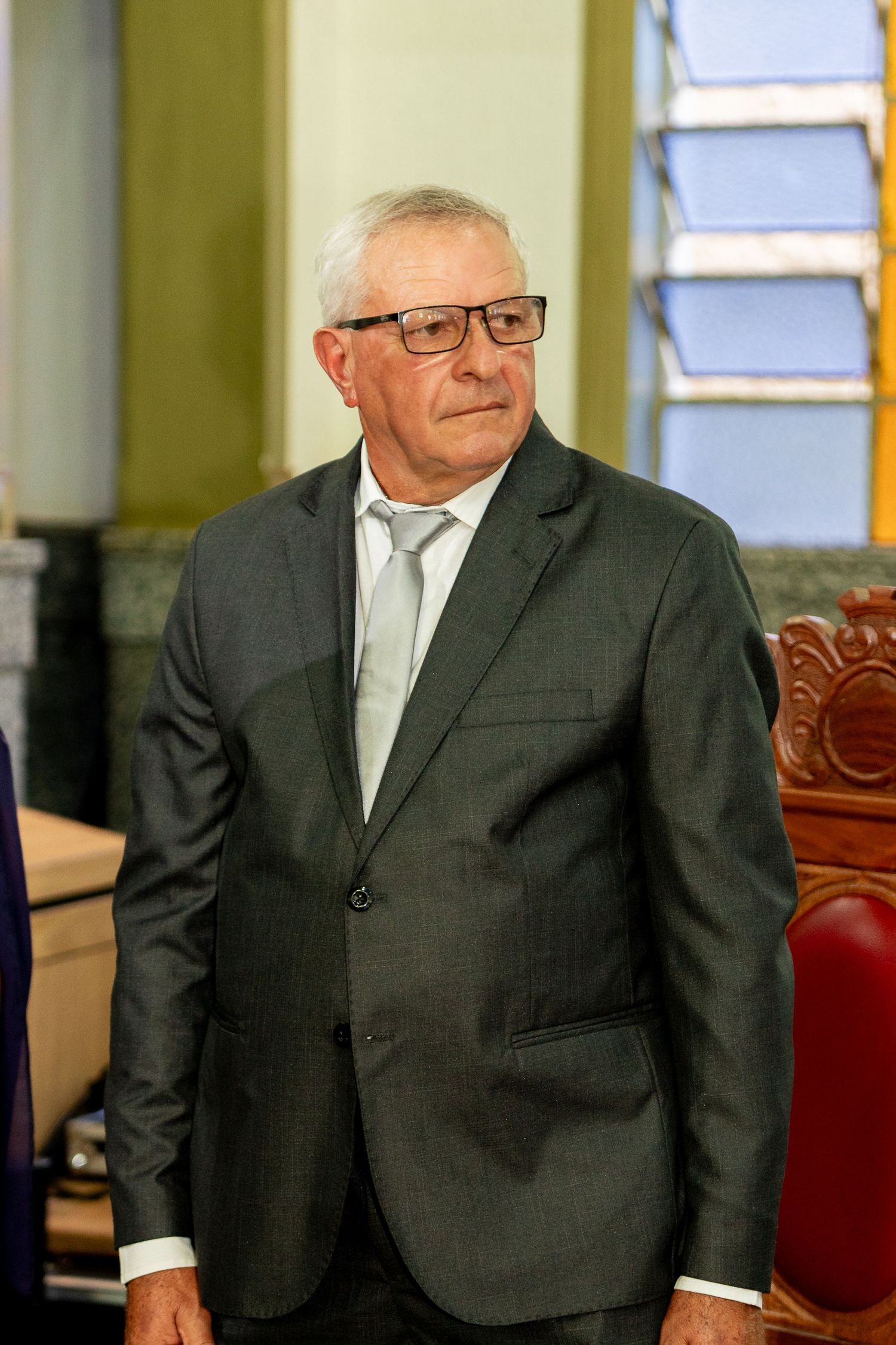 Elderly man in gray suit and glasses standing in formal indoor setting.