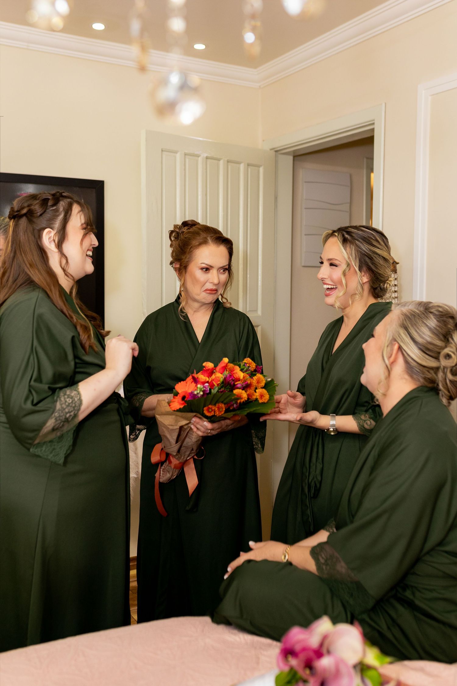 Four bridesmaids in matching dark green robes gather together, one holding a vibrant bouquet of colorful flowers.