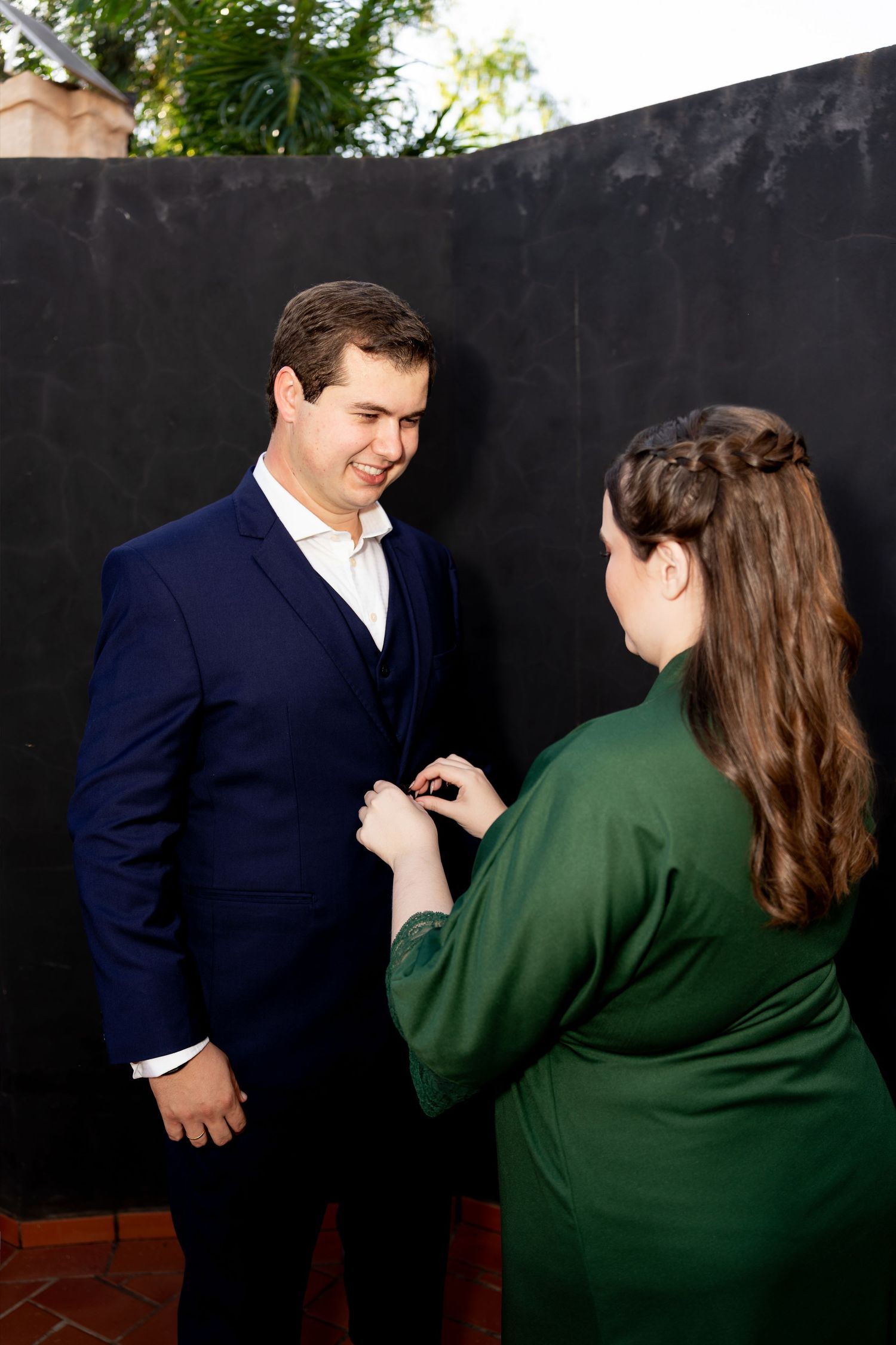 Woman in green dress pinning boutonniere on man in navy blue suit against dark wall outdoors.