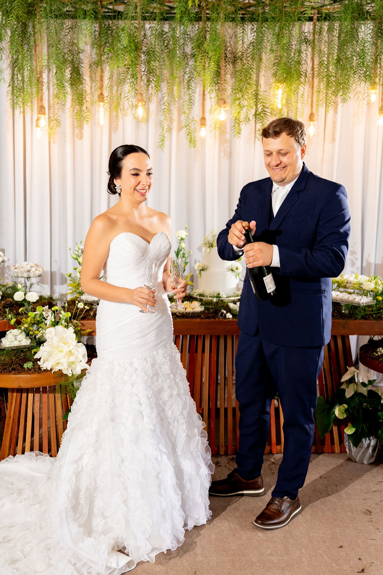 Bride and groom opening champagne bottle at wedding reception with floral decor and hanging lights.