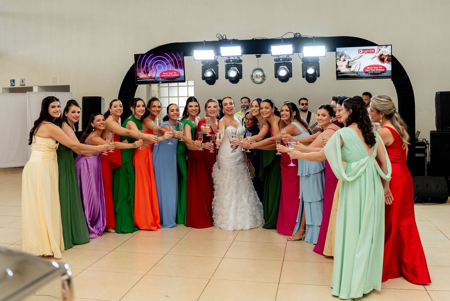 Bride in white gown surrounded by bridesmaids in colorful rainbow dresses holding hands in a circle at wedding reception.