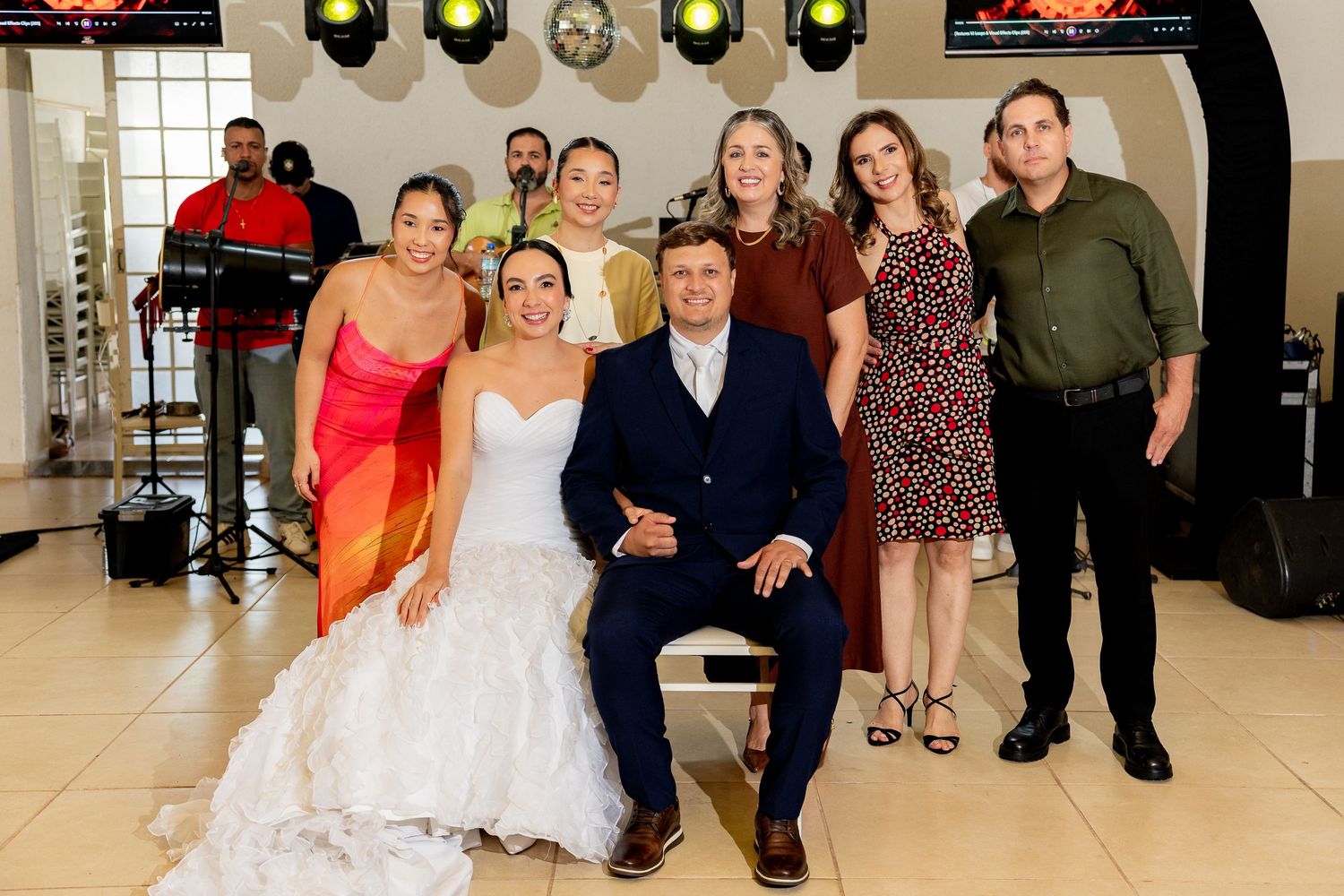 Wedding couple seated with family and friends posing together at a reception venue with stage lighting.