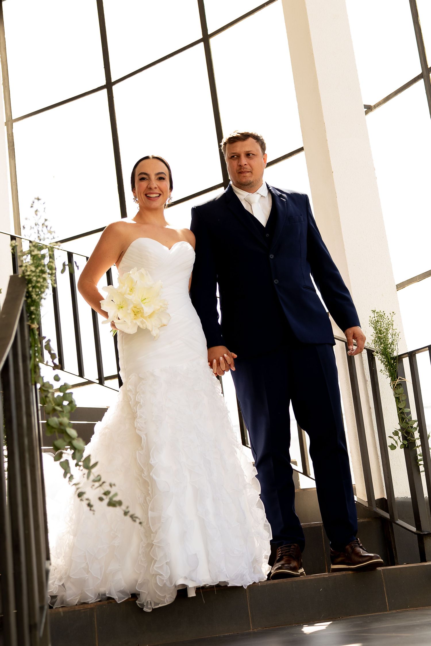 Bride in white ruffled gown holding bouquet stands with groom in navy suit on staircase at wedding venue.