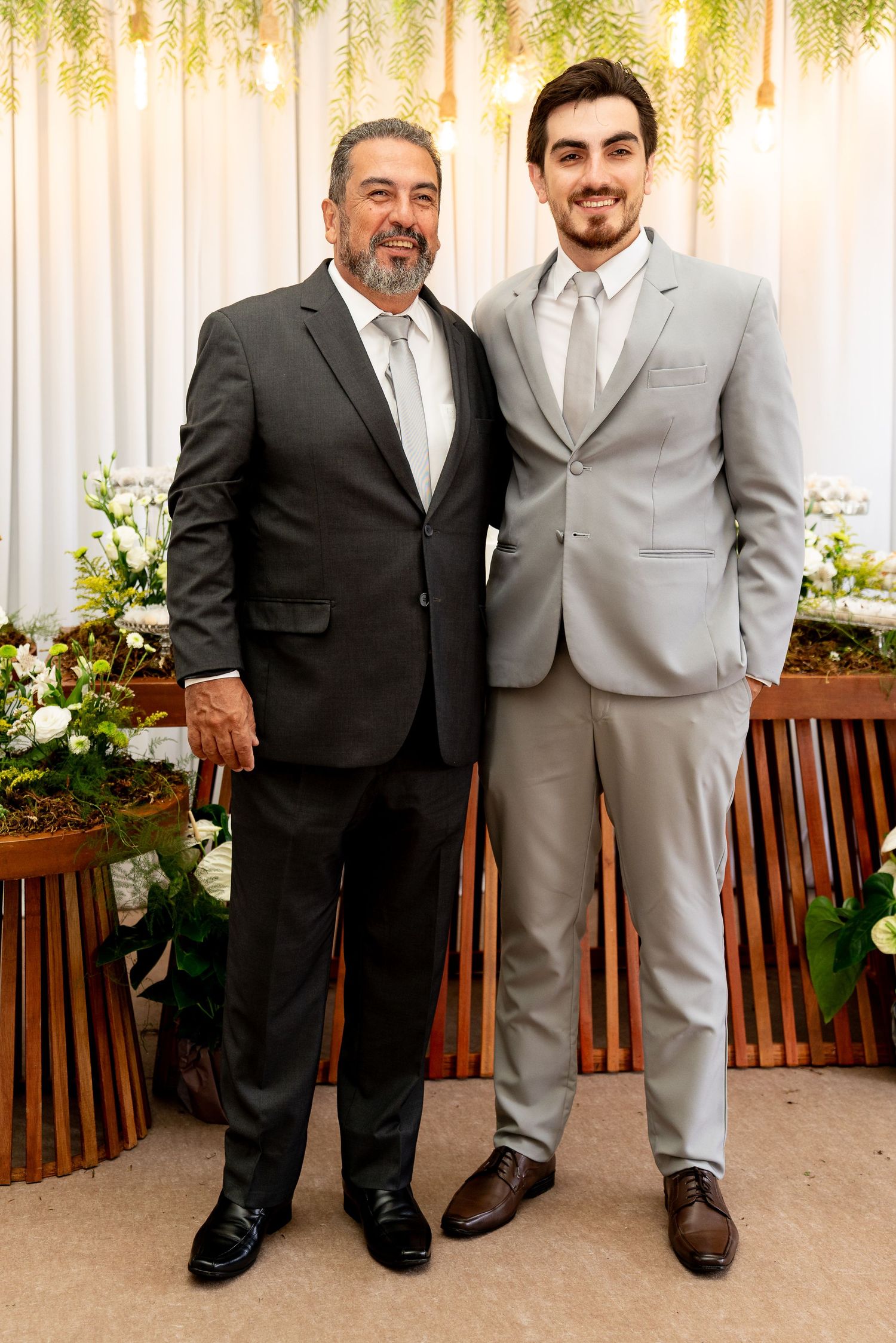 Two men in suits pose together at a wedding venue decorated with white flowers and greenery.