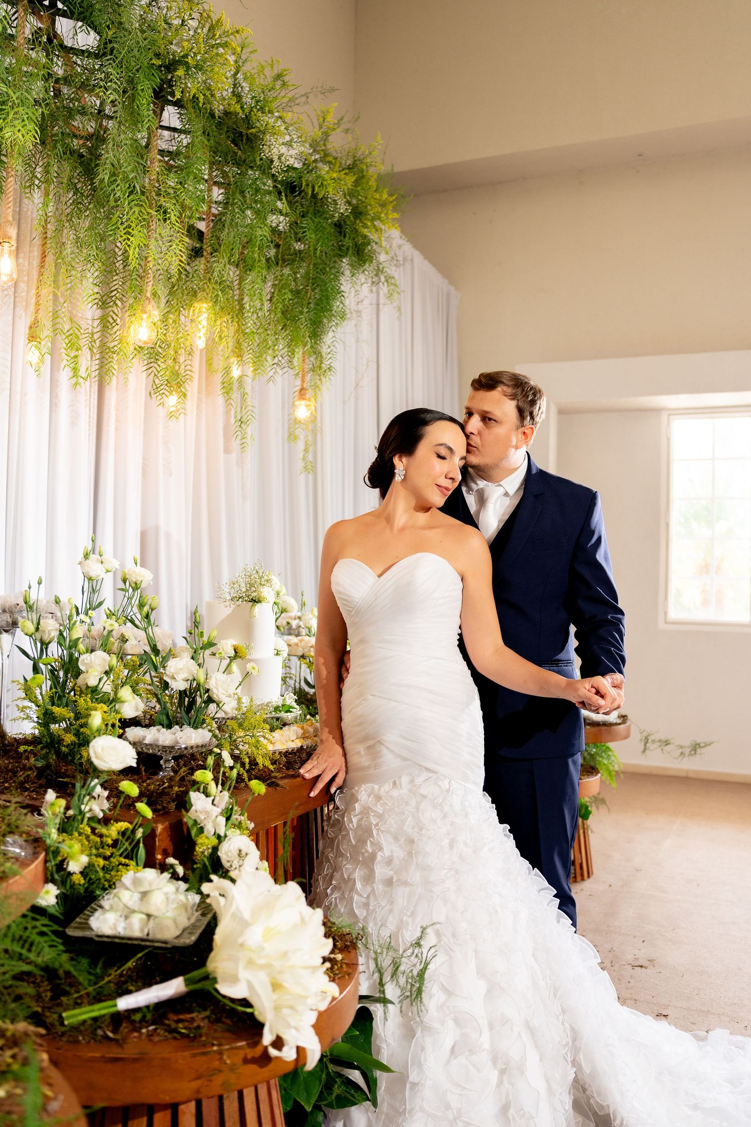 Bride in white mermaid gown and groom in navy suit pose by floral arrangements under hanging greenery and lights.