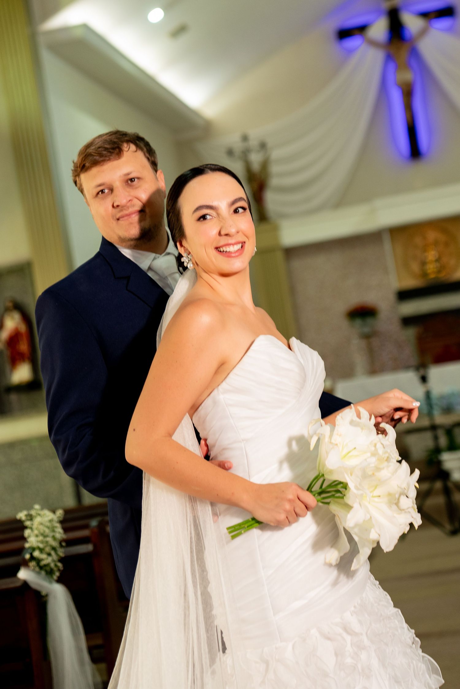 A smiling bride in a white strapless gown holds a white bouquet as her groom in a navy suit stands behind her in a church.