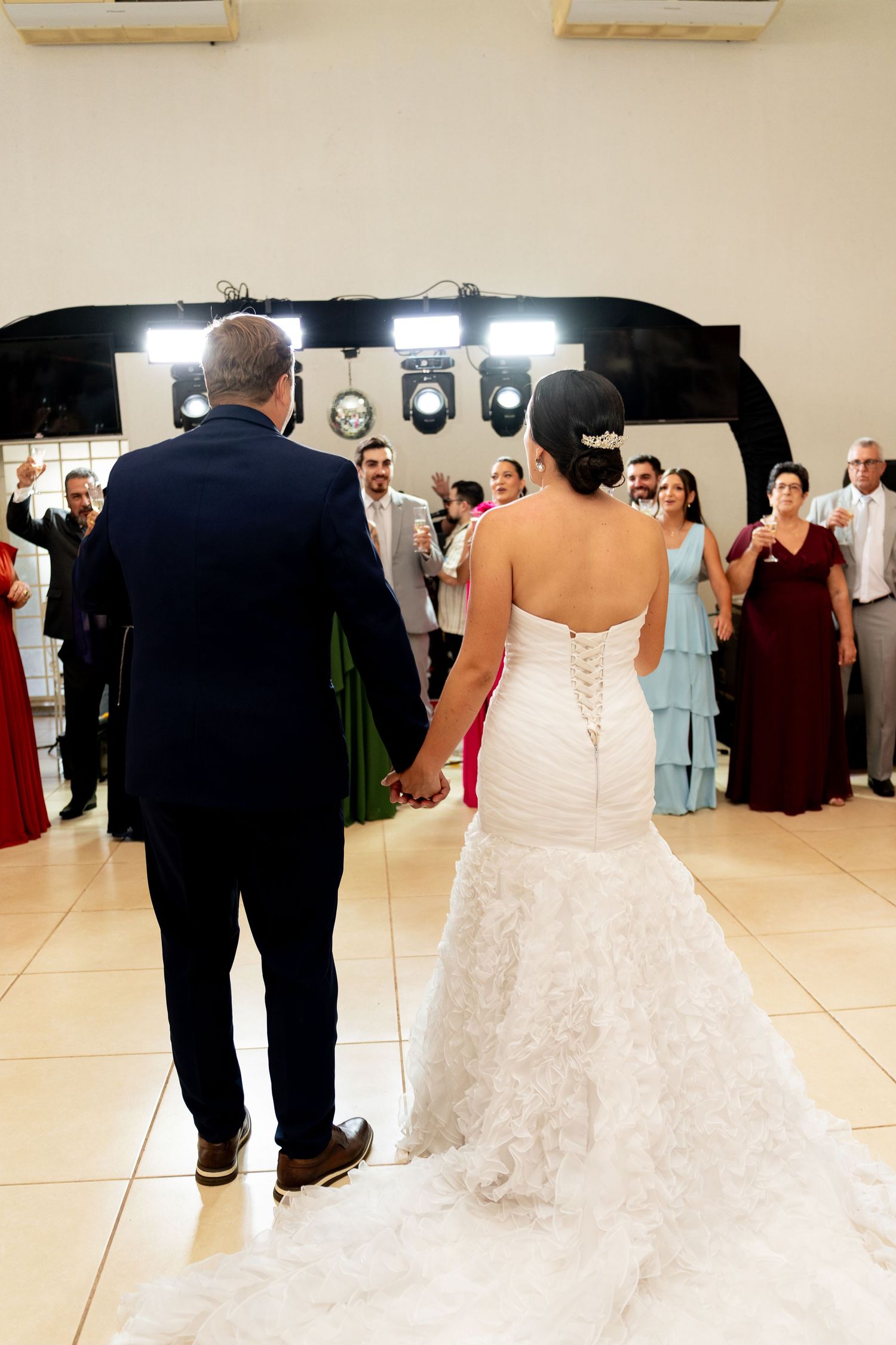 Bride in white lace gown and groom in navy suit hold hands entering wedding reception as guests applaud.