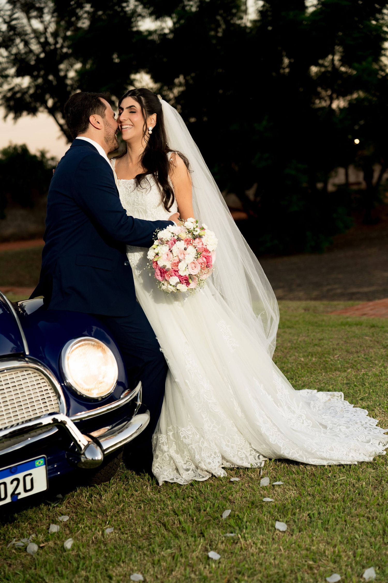 Bride and groom sharing a kiss beside a vintage blue car, bride holding a pink and white bouquet in a lace wedding gown.
