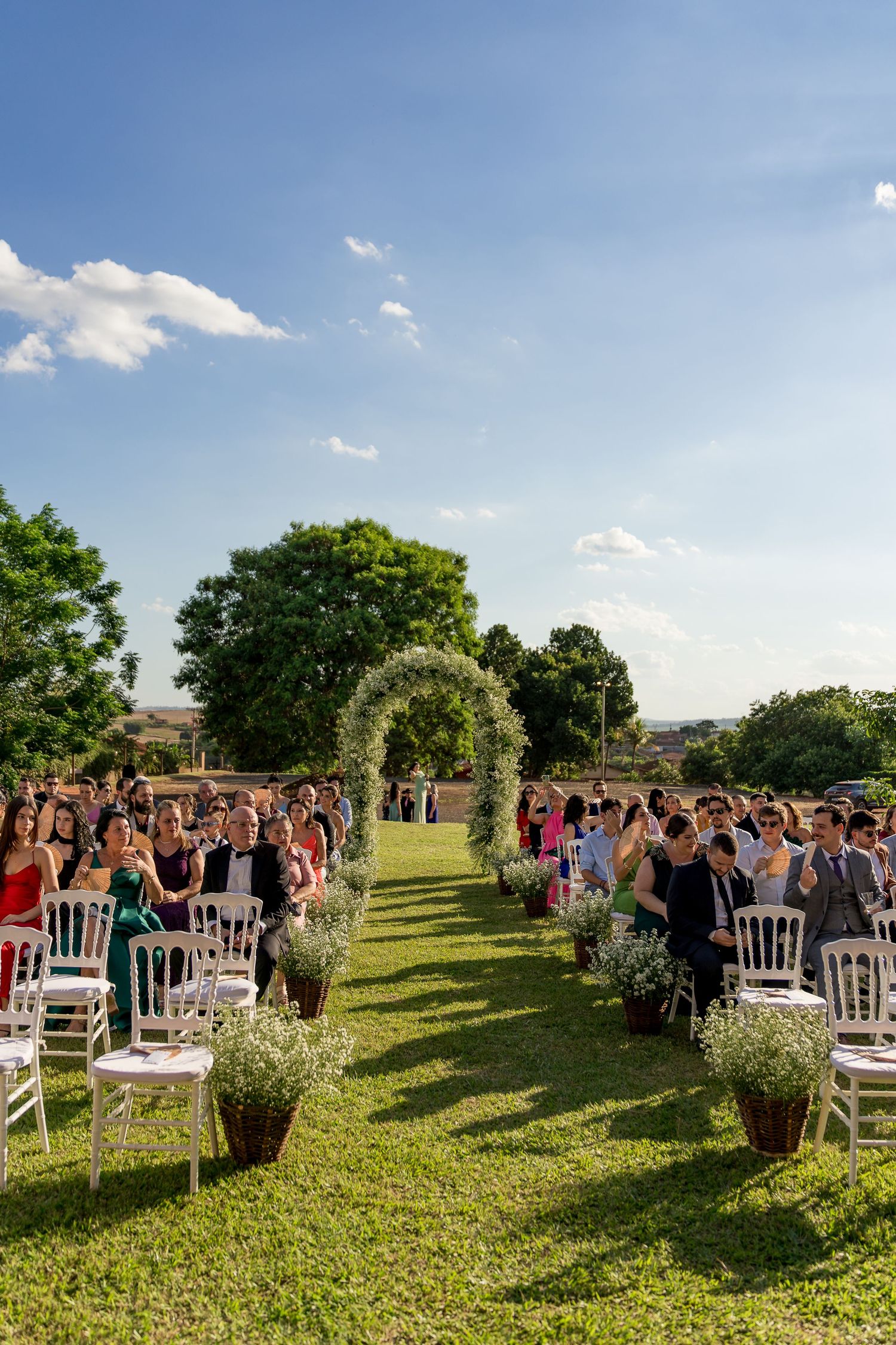 Elegant outdoor wedding ceremony with white chairs, floral arch, and guests seated on a green lawn under blue sky.