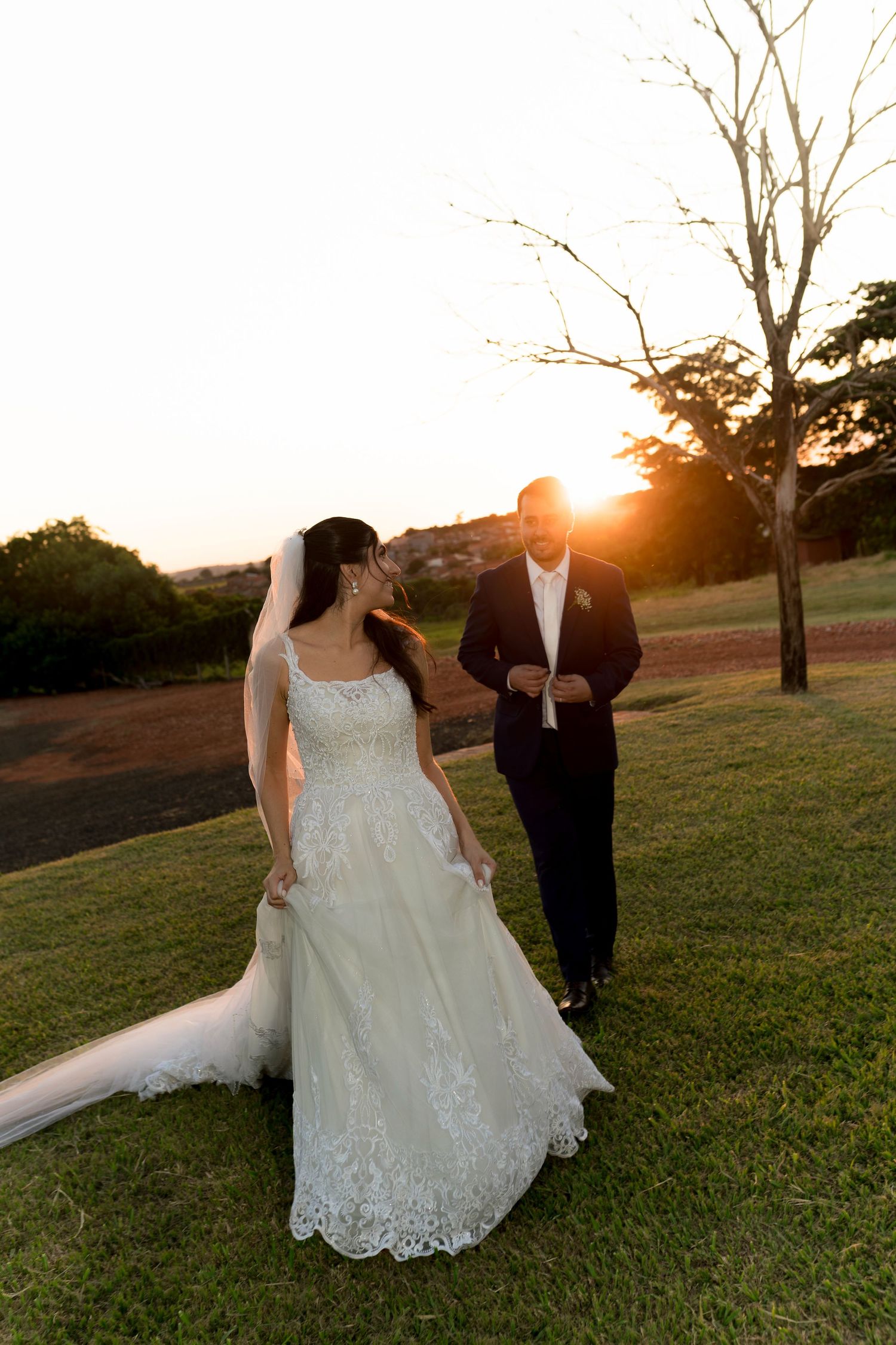 Bride in lace gown and groom in tuxedo walk together on green lawn at golden sunset during outdoor wedding.