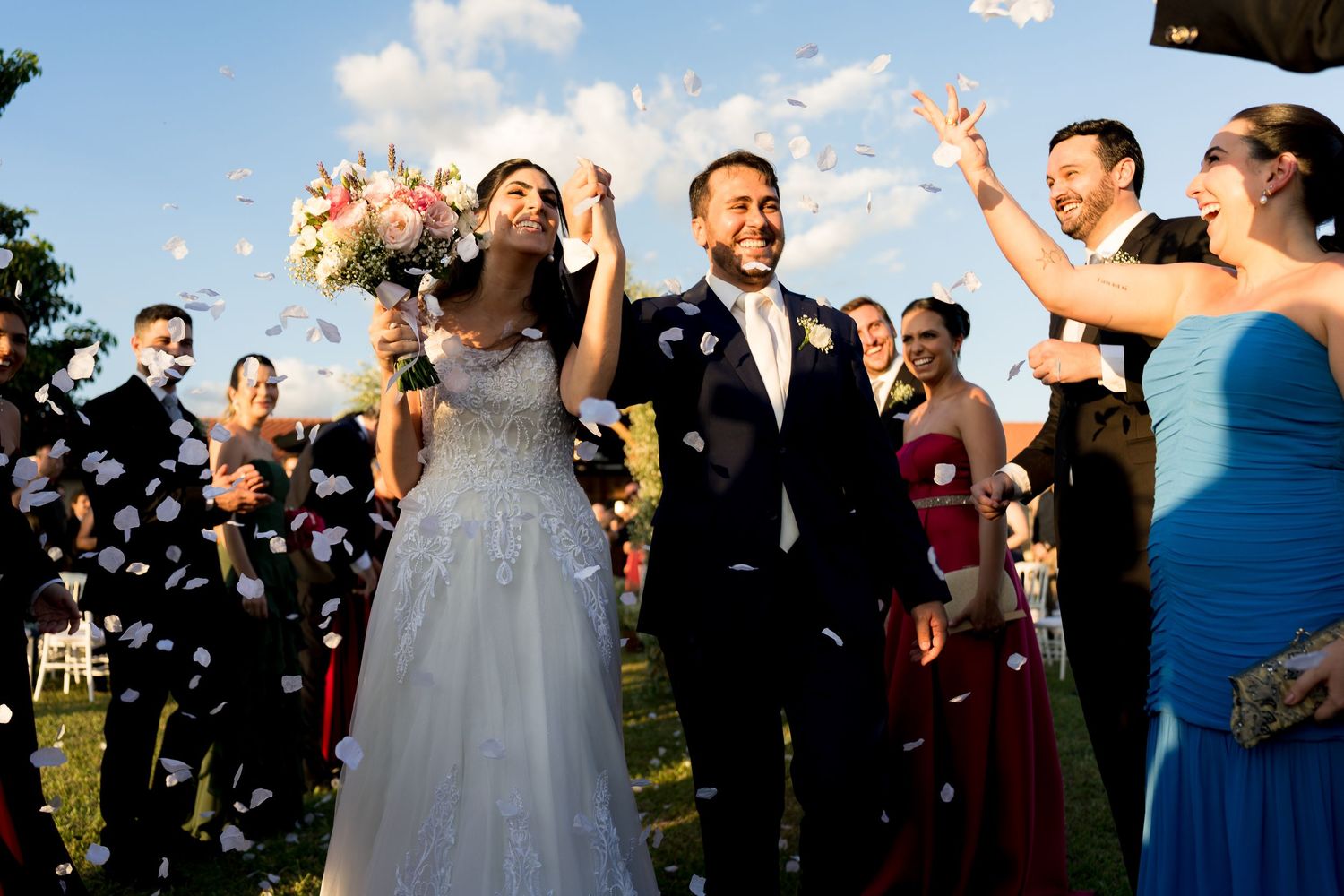 Bride and groom walk through confetti shower surrounded by smiling wedding guests outdoors on a sunny day.