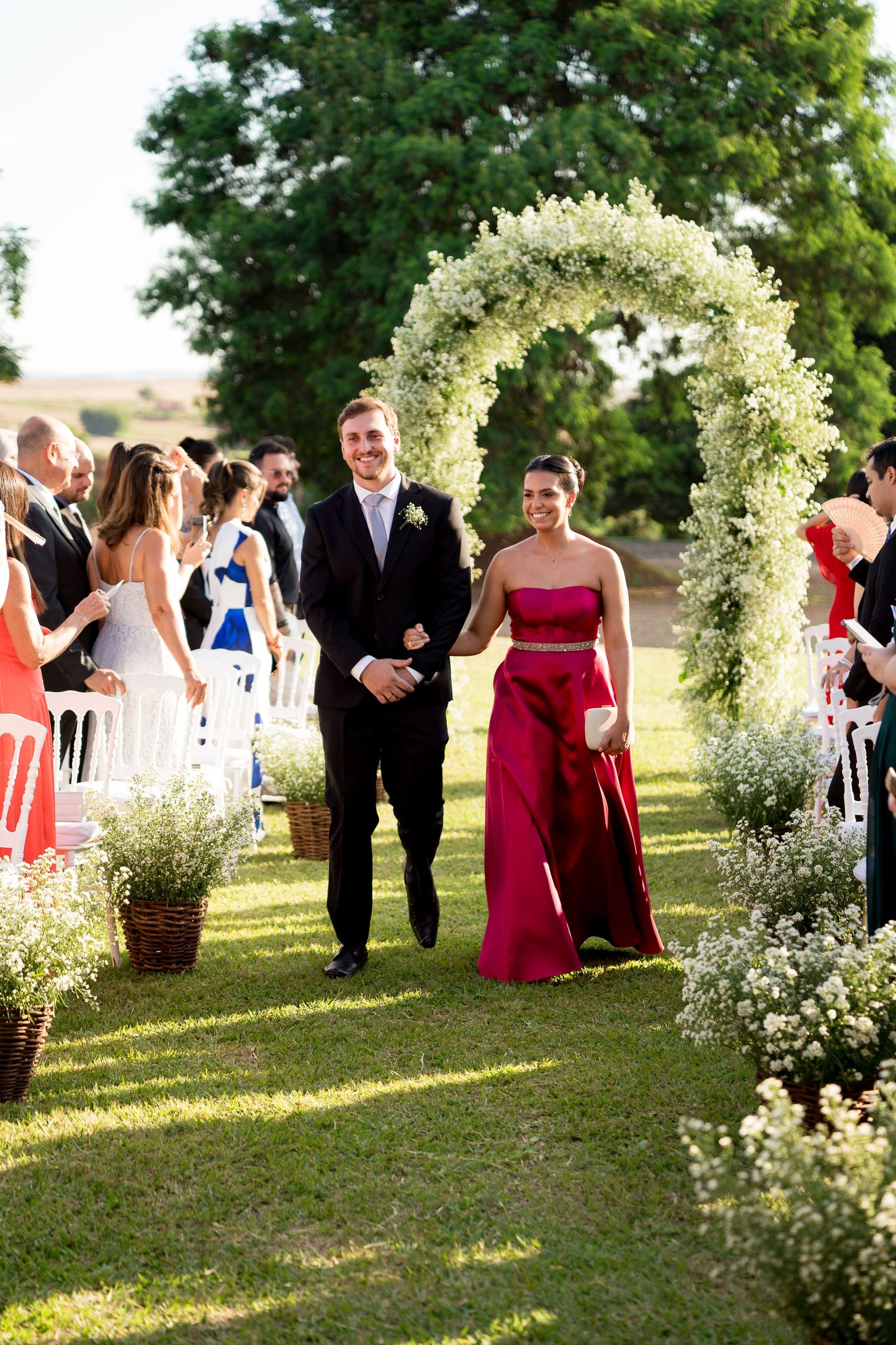 Couple walks down outdoor wedding aisle under floral arch, guests applaud, woman in burgundy gown, man in dark suit.