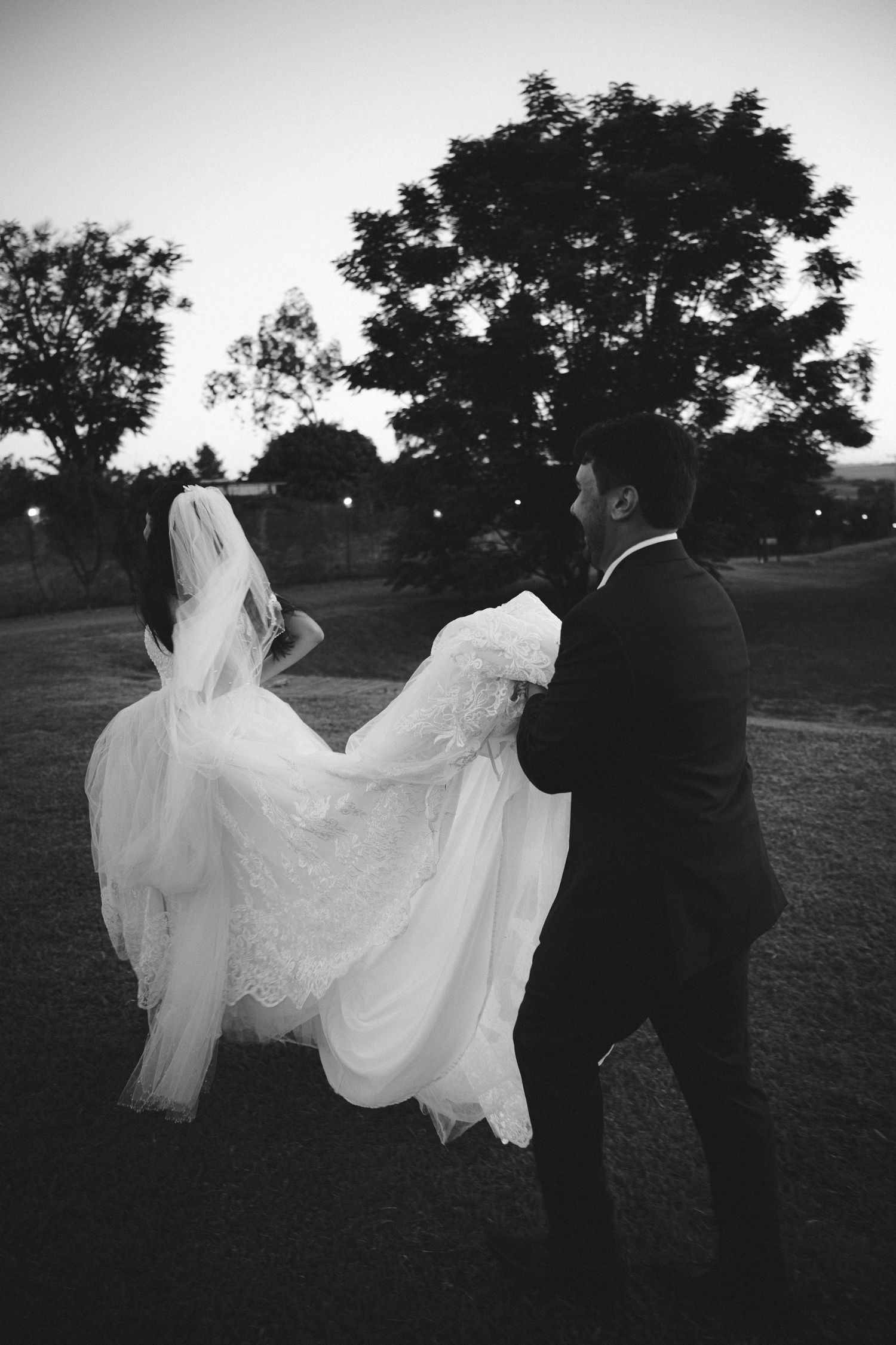 Black and white photo of a groom helping his bride with her flowing lace wedding dress and veil at dusk outdoors.